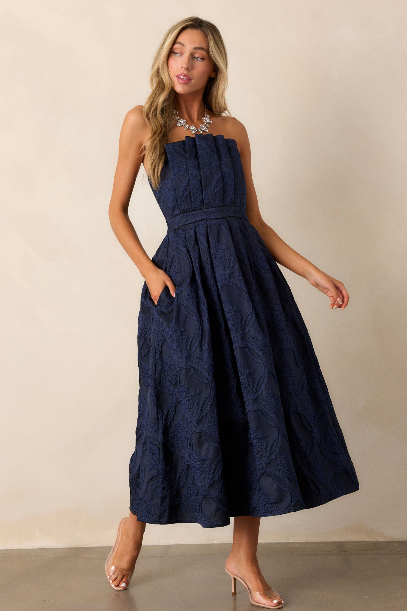 Side view of a navy midi dress featuring a pleated bodice, a slightly pleated skirt, a jacquard design, functional hip pockets, a smocked back, a discrete back zipper and a strapless design.