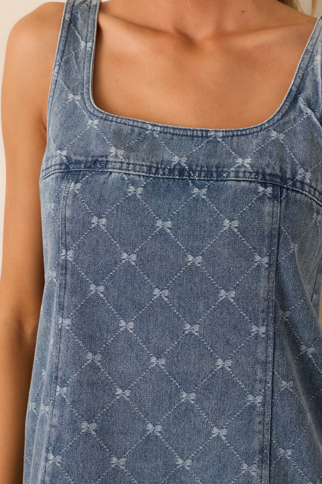 Detailed view of the argyle bow design, the square neckline, and sleeveless straps on this denim shift mini dress.