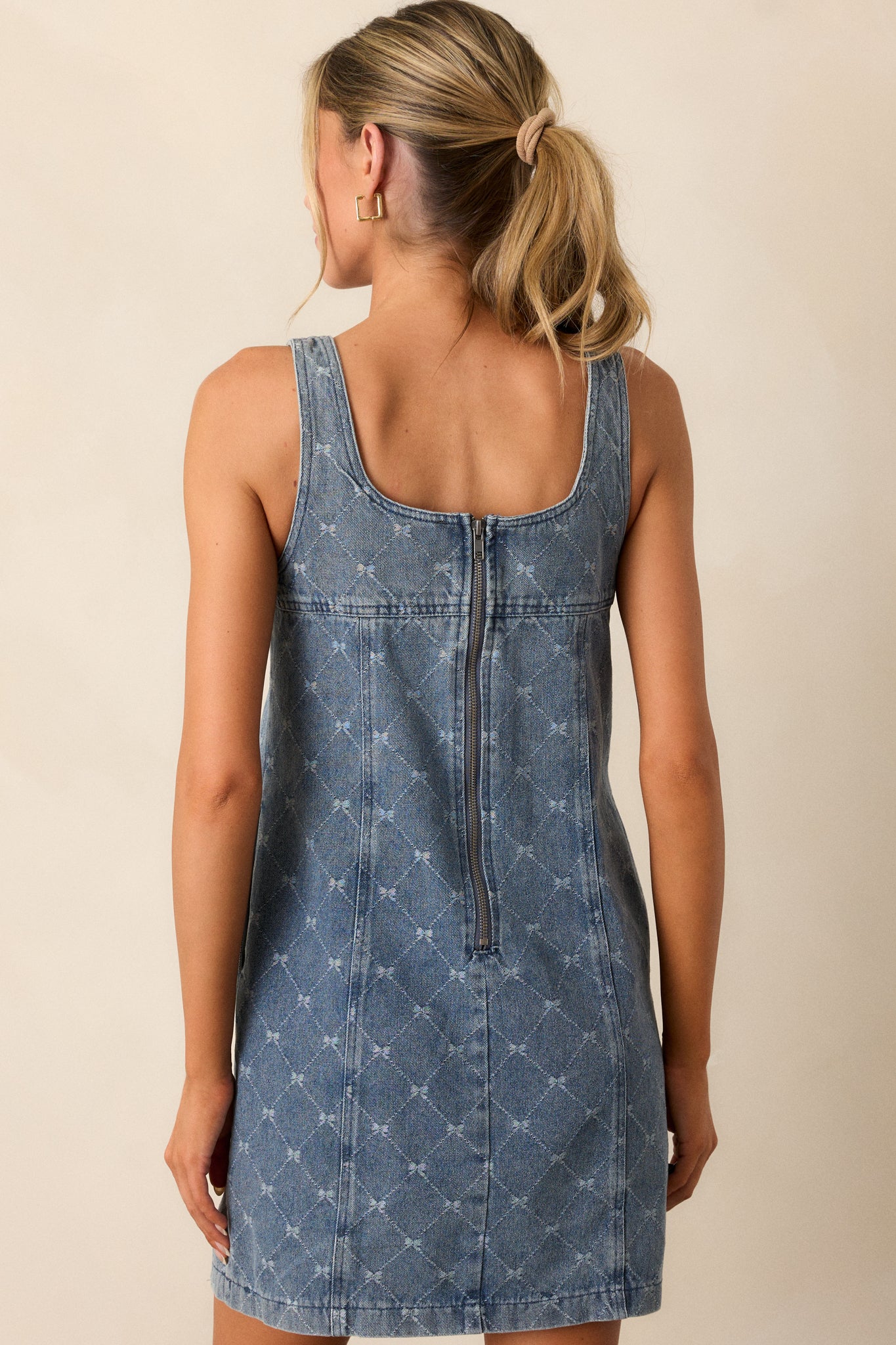 Back view of a medium wash mini dress featuring a square neckline, an argyle bow print, a shift silhouette, a denim cotton blend fabric, and a sleeveless design.