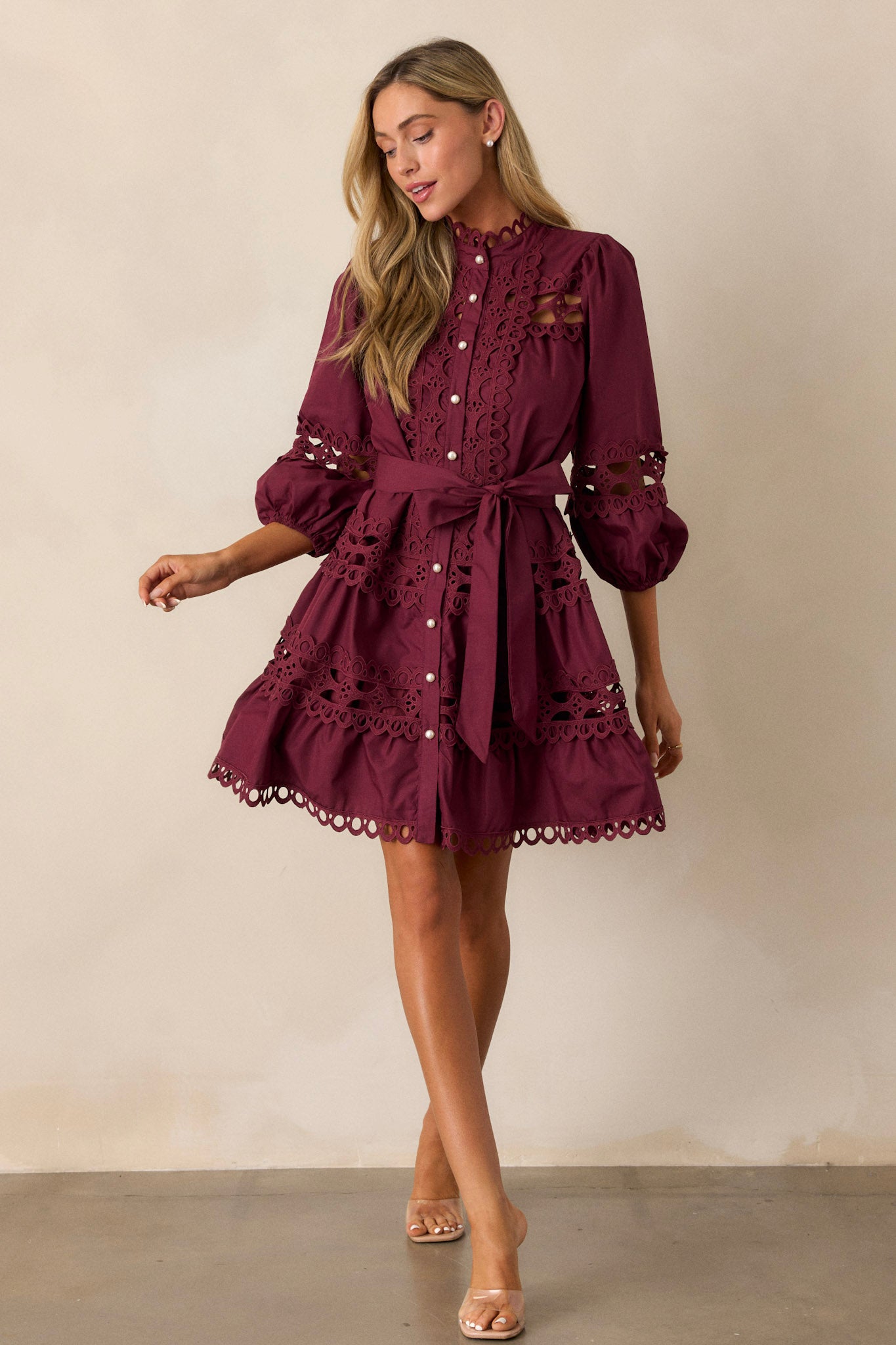 Full view of this  plum mini dress featuring a high neckline, a functional button front with faux pearl buttons, an optional self-tie belt, elastic cuffed 3/4 length sleeves, and eyelet detailing throughout.