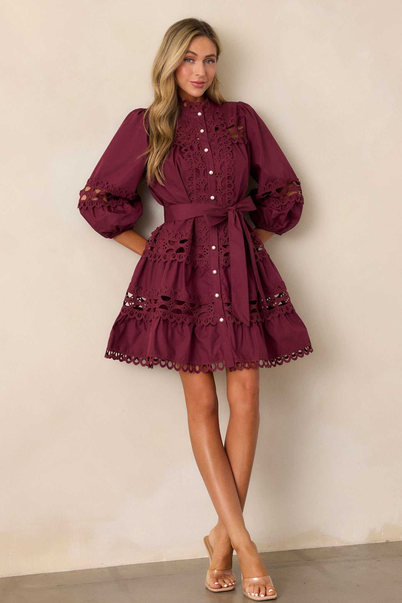 Angled view of this  plum mini dress featuring a high neckline, a functional button front with faux pearl buttons, an optional self-tie belt, elastic cuffed 3/4 length sleeves, and eyelet detailing throughout.