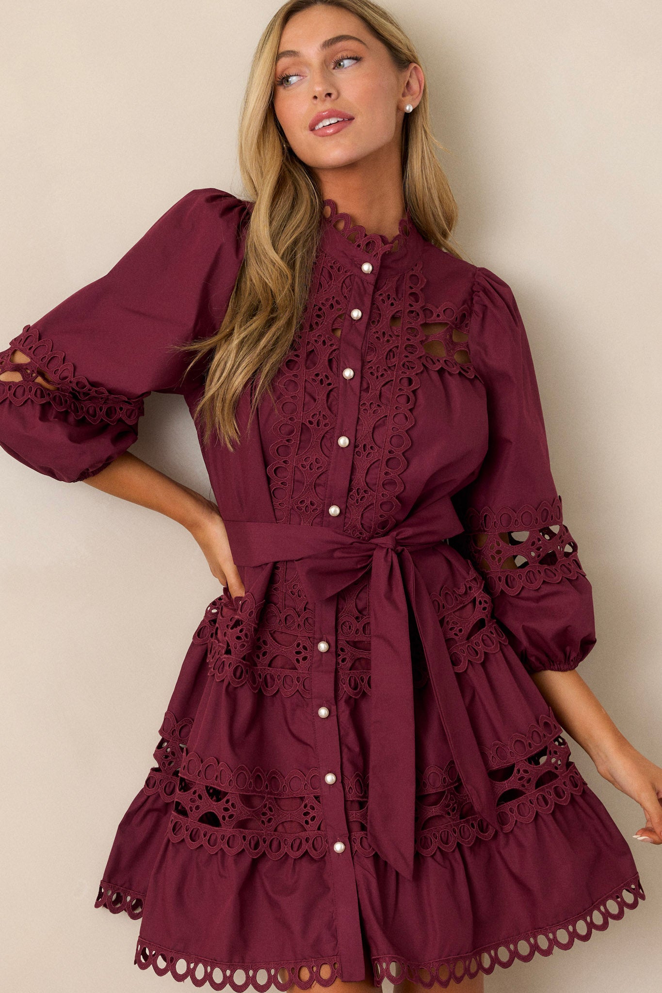 This plum mini dress features a high neckline, a functional button front with faux pearl buttons, an optional self-tie belt, elastic cuffed 3/4 length sleeves, and eyelet detailing throughout.