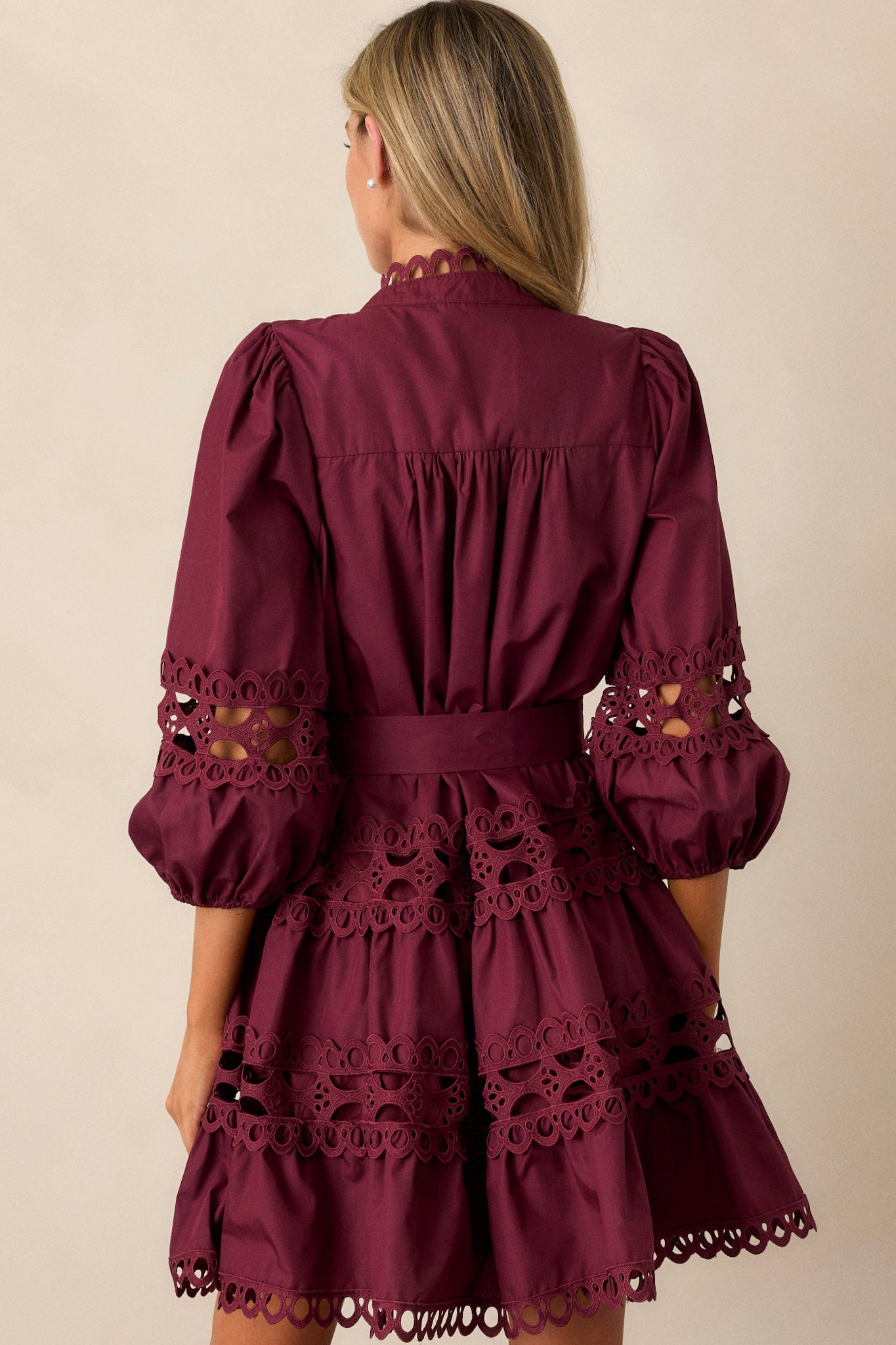 Back view of this  plum mini dress featuring a high neckline, a functional button front with faux pearl buttons, an optional self-tie belt, elastic cuffed 3/4 length sleeves, and eyelet detailing throughout.