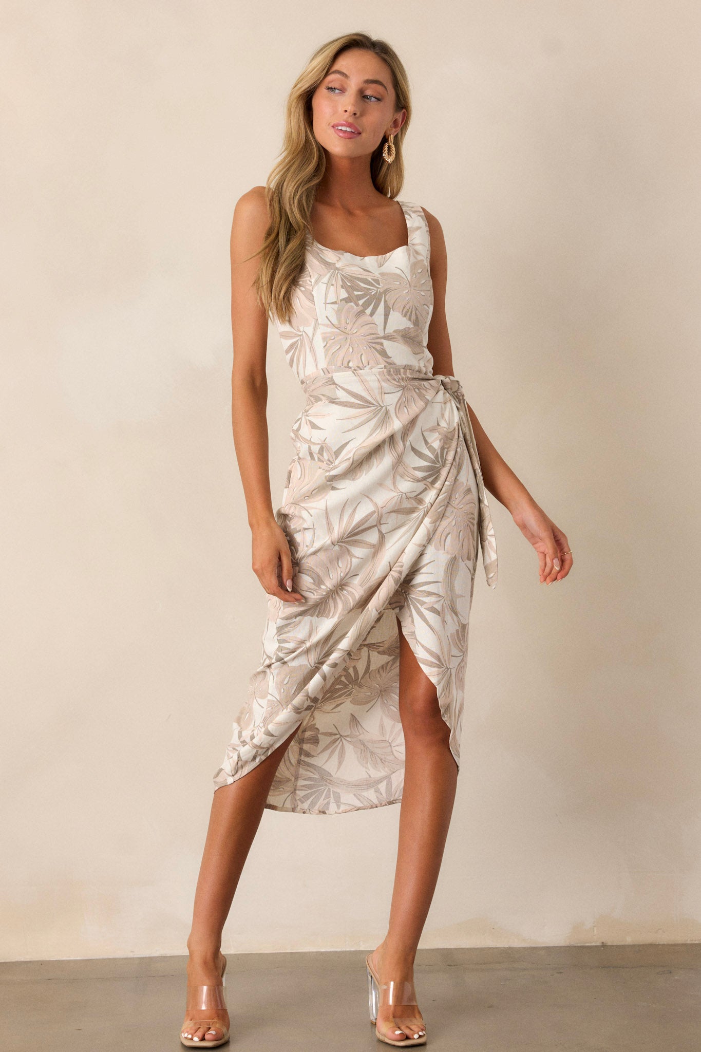 Front view of this taupe midi dress that features a square neckline, thick straps, a smocked back insert, a discrete zipper, a self-tie wrap feature, and a neutral tropical print.