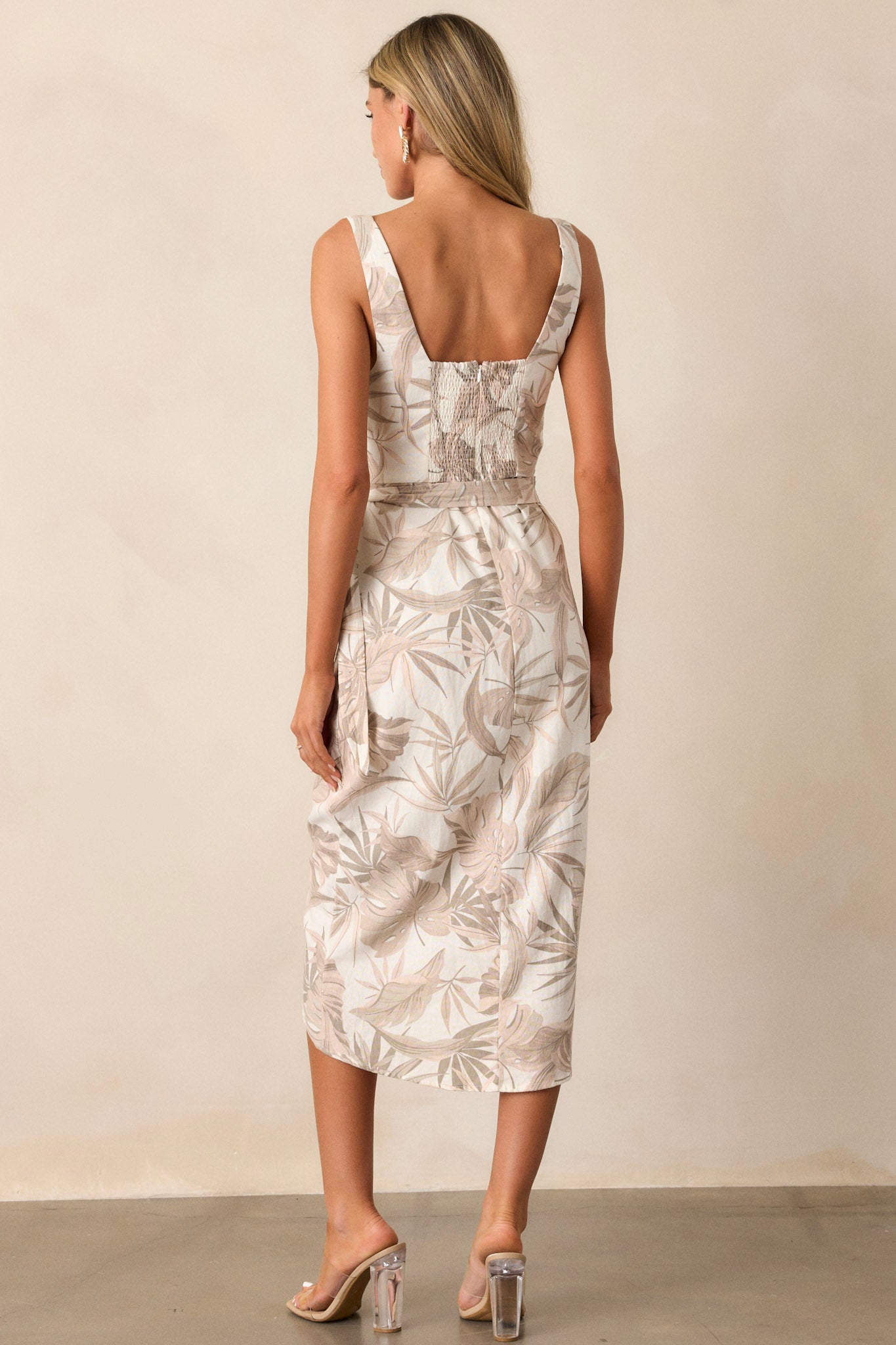 Back view of this taupe midi dress that features a square neckline, thick straps, a smocked back insert, a discrete zipper, a self-tie wrap feature, and a neutral tropical print.