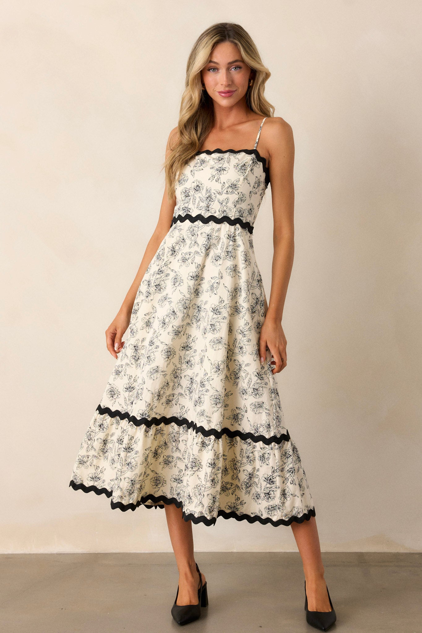 Front view of an ivory maxi dress featuring a square neckline, black ruffle trim, a tiered design, a black floral print, a discrete back zipper, and adjustable spaghetti straps.