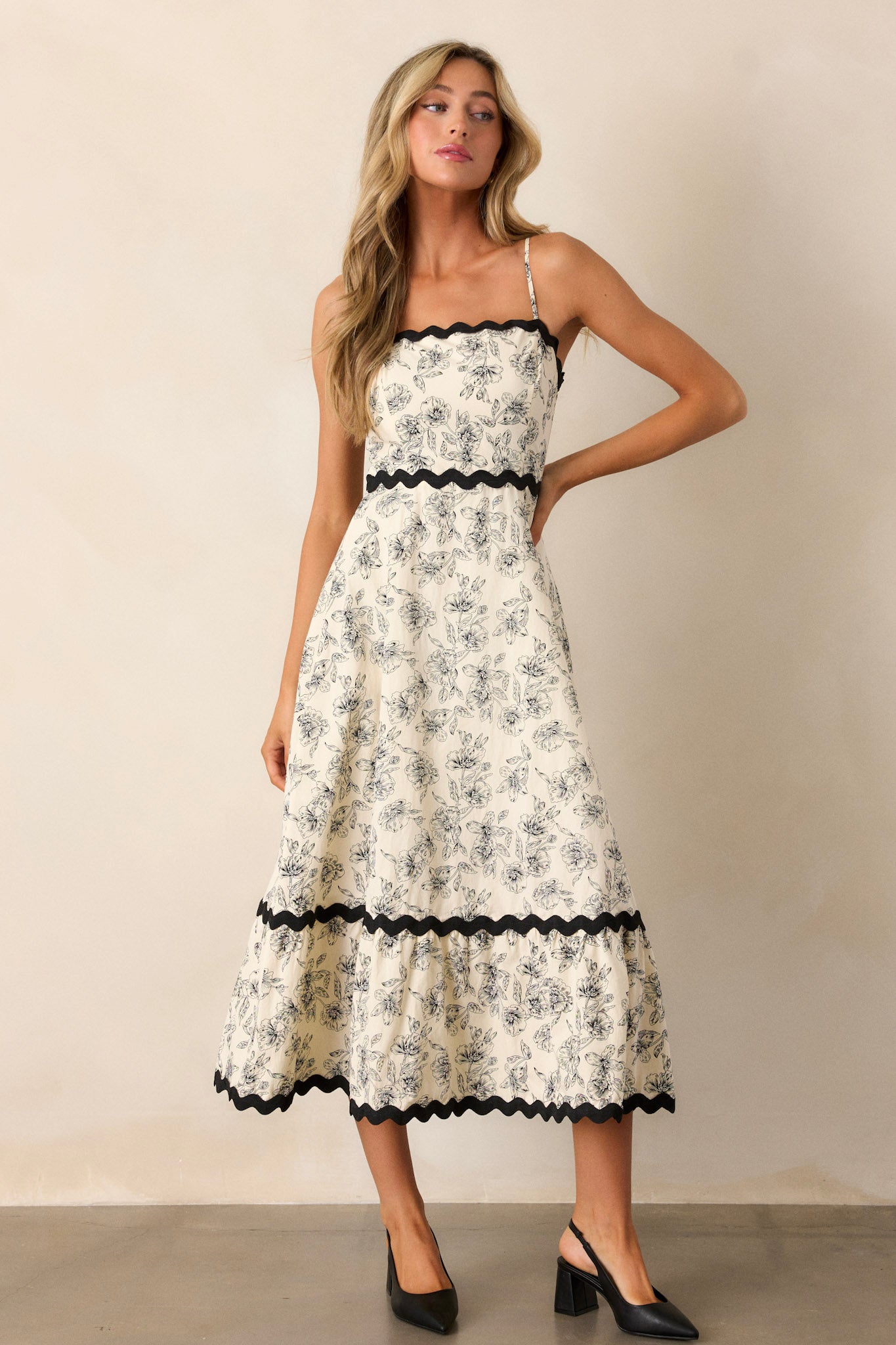 Full length view of an ivory maxi dress featuring a square neckline, black ruffle trim, a tiered design, a black floral print, a discrete back zipper, and adjustable spaghetti straps