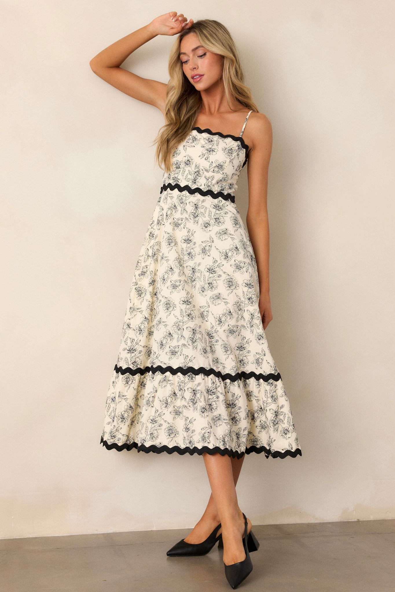 Angled front view of an ivory maxi dress featuring a square neckline, black ruffle trim, a tiered design, a black floral print, a discrete back zipper, and adjustable spaghetti straps.