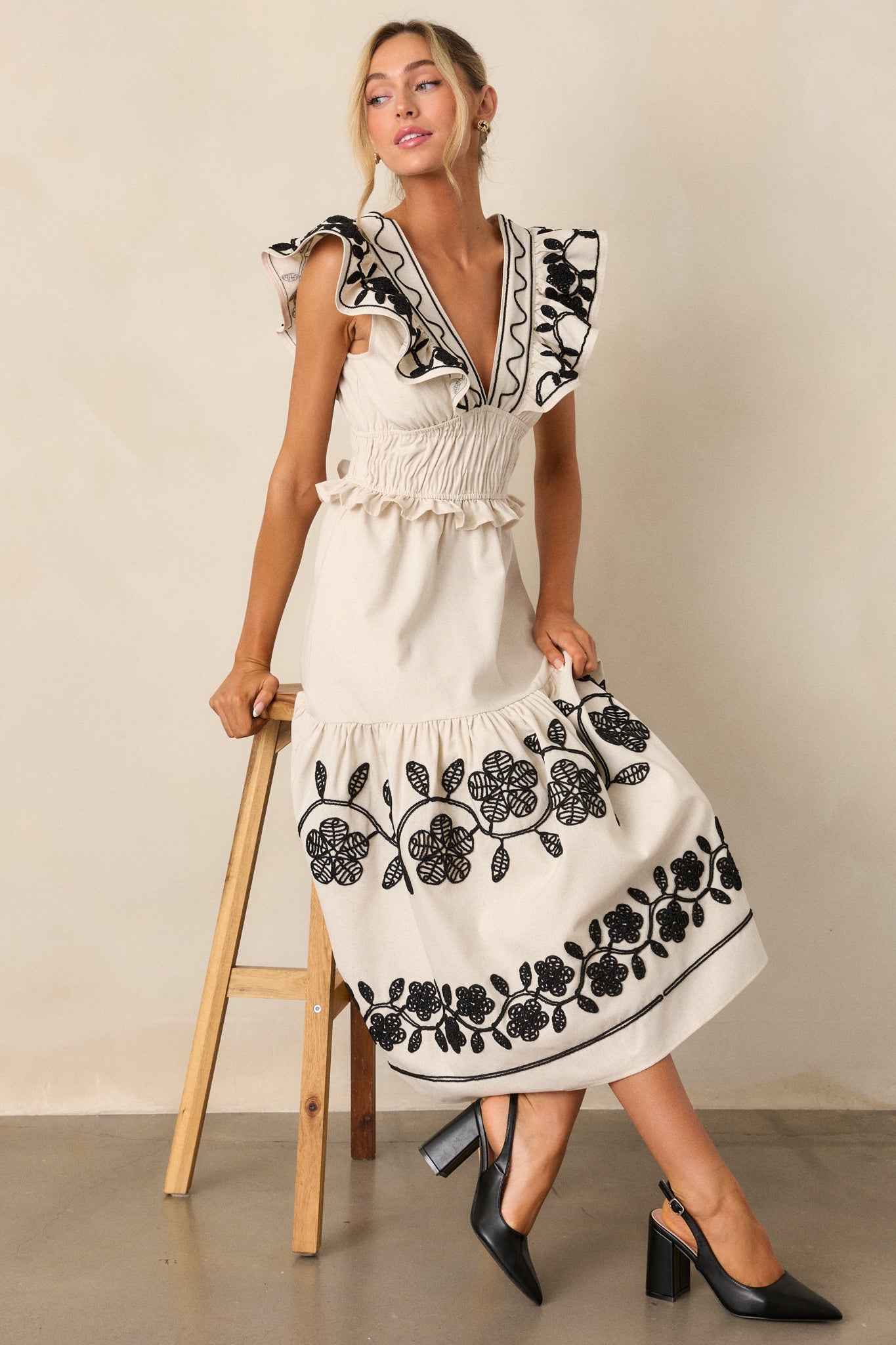 Seated full-length view of a model wearing the beige dress, showing the plunging v-neckline, smocked waist, functional hip pockets, short flutter sleeves, and embroidered detailing throughout.