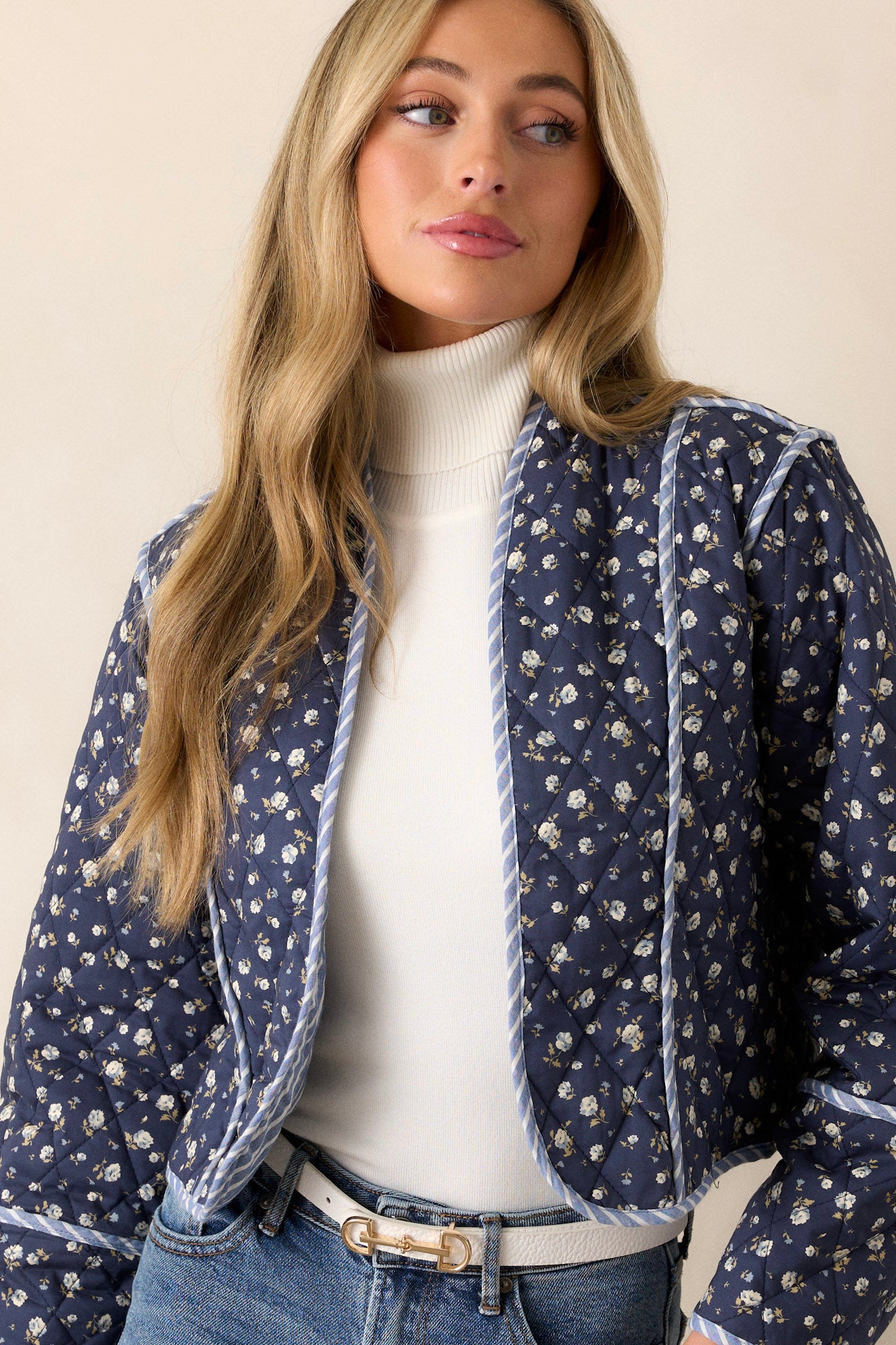 Front angled view of this navy jacket that features an open front, a rounded neckline, a small floral pattern, a blue and white striped interior, a quilted material, a blue and white striped trim and long sleeves.