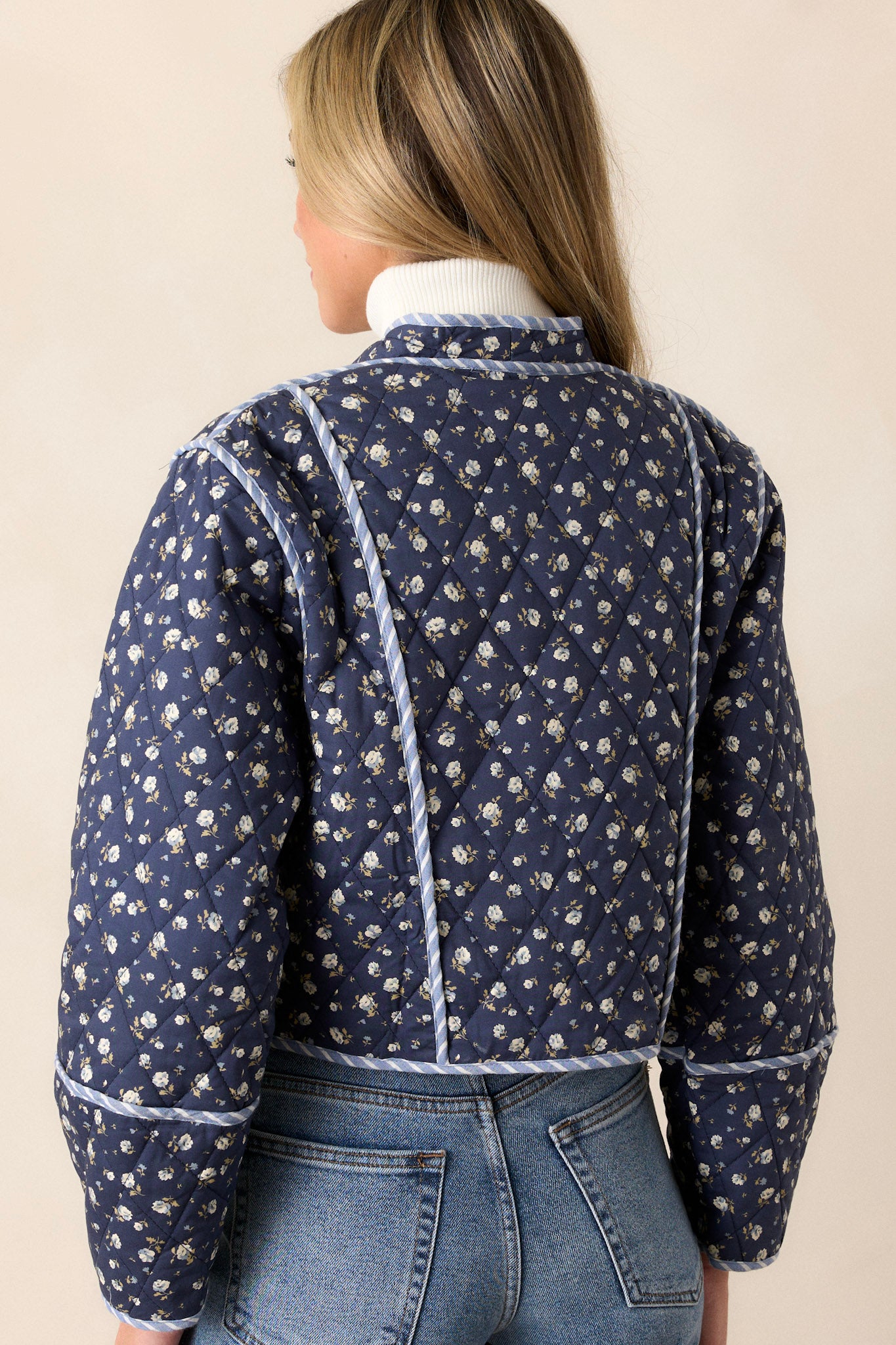 Back view of this navy jacket that features an open front, a rounded neckline, a small floral pattern, a blue and white striped interior, a quilted material, a blue and white striped trim and long sleeves.