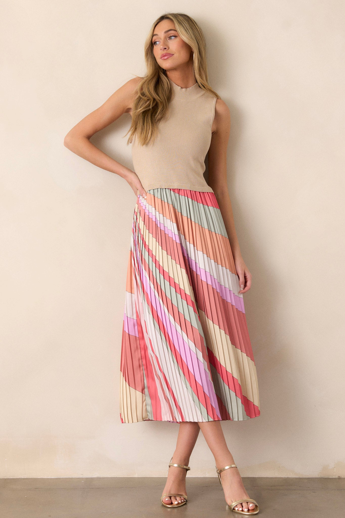 Front angled view of a taupe midi dress featuring a high neckline, a ribbed sweater-like bodice, a multi-color pleated skirt, a sleeveless design, and a flowing silhouette