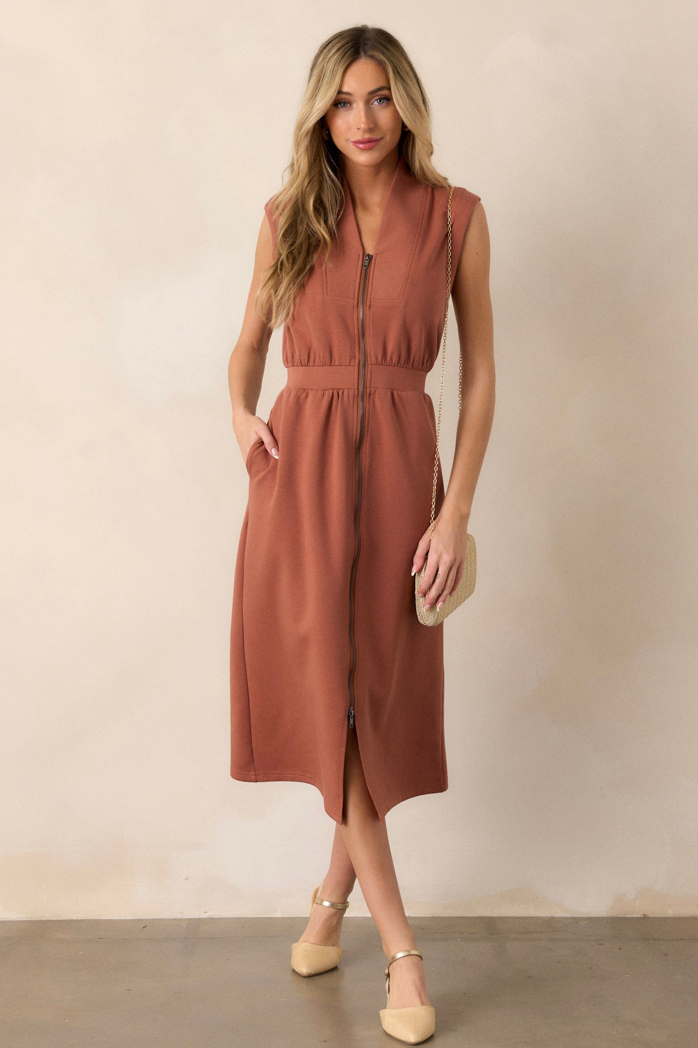 Full body of this terracotta dress that features a v-neckline, a slight collar, a functional full zip front, functional hip pockets, and a sleeveless design.