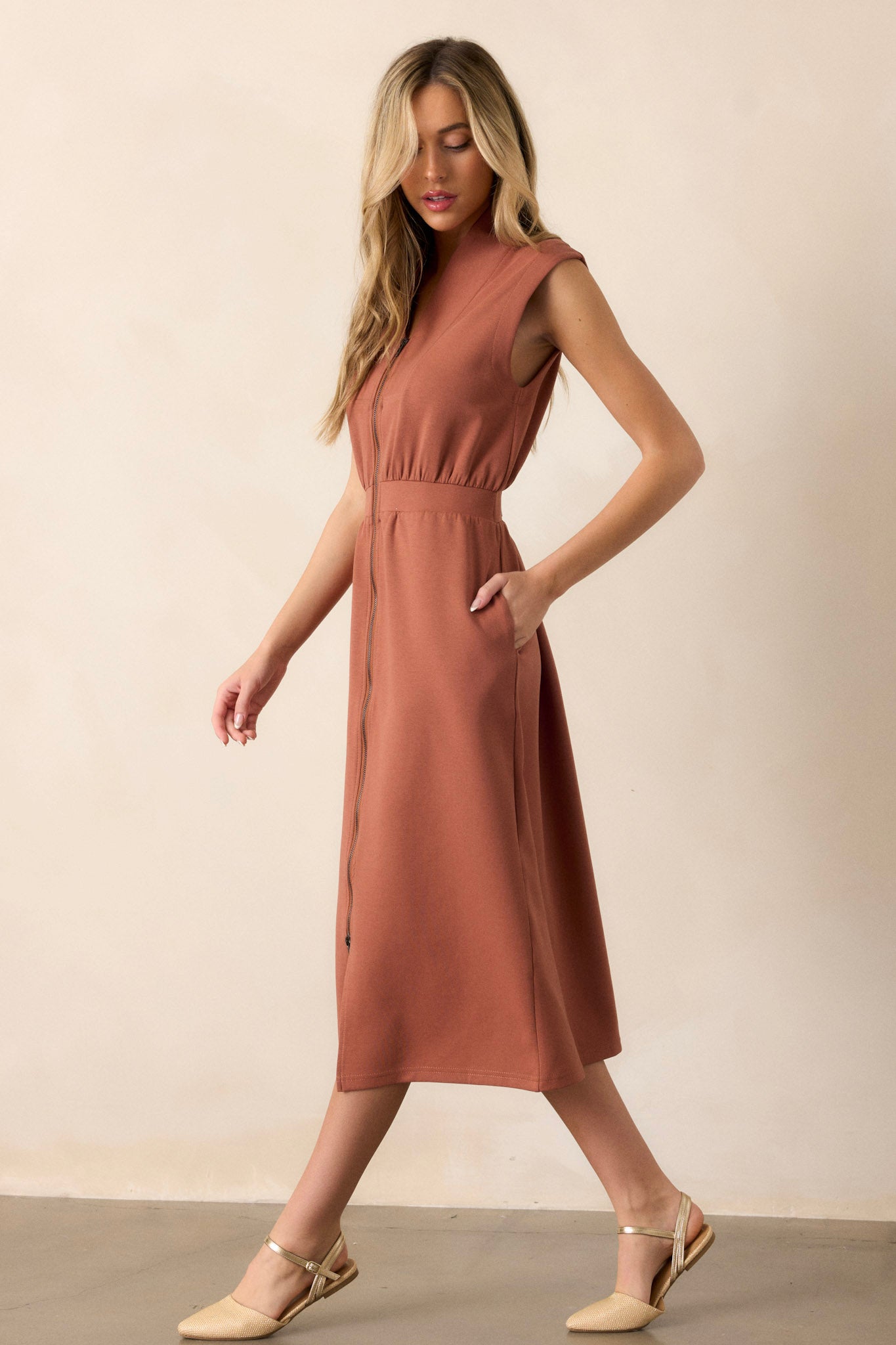 Side view of this terracotta dress that features a v-neckline, a slight collar, a functional full zip front, functional hip pockets, and a sleeveless design.