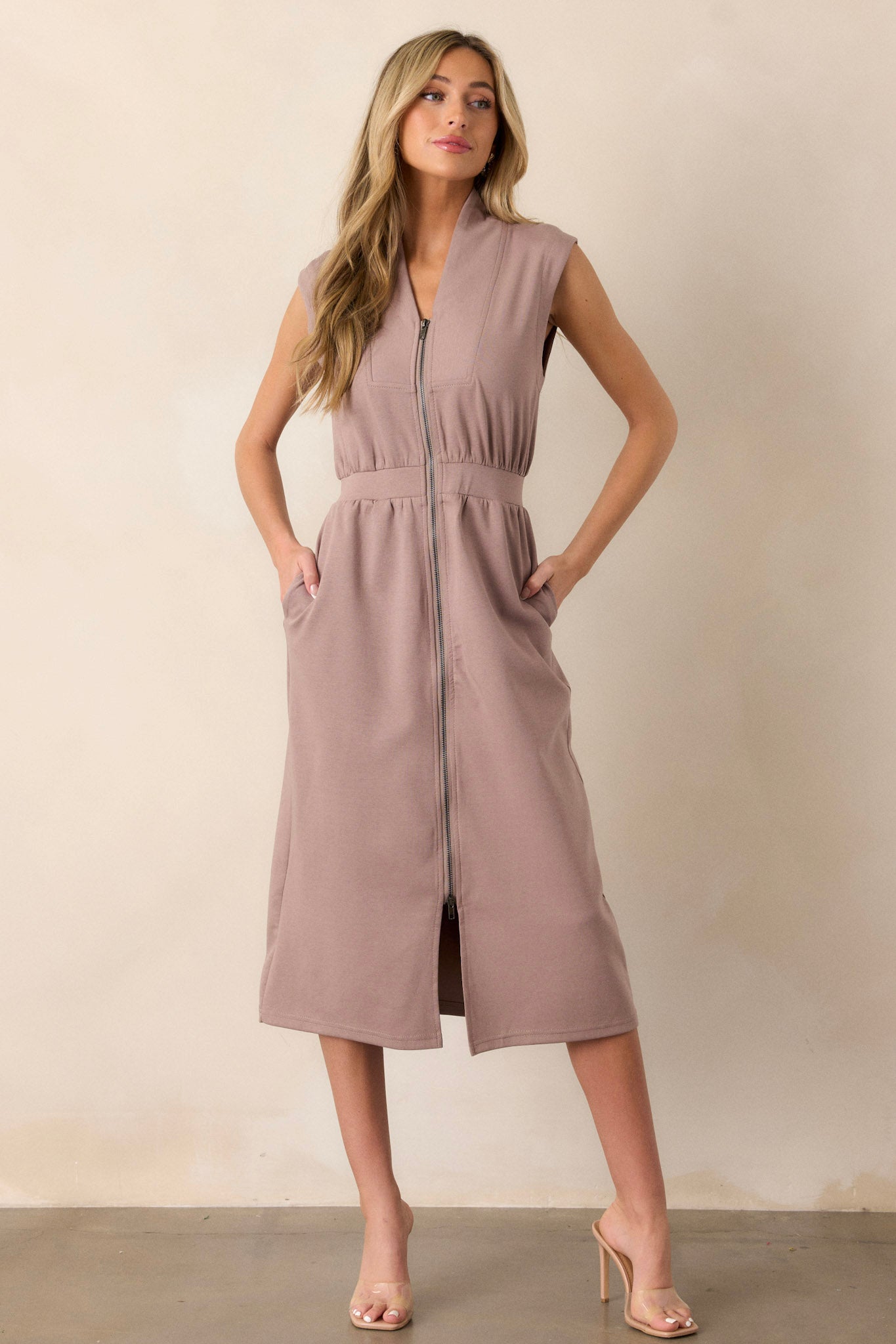 Front angled  view of this taupe dress that features a v-neckline, a slight collar, a functional zip front, functional hip pockets, and an elastic waistband.