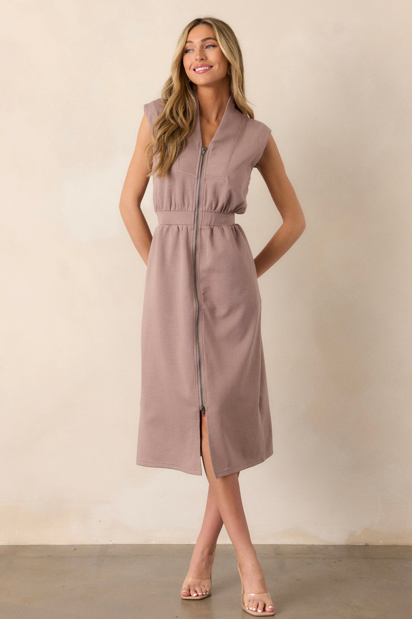 Angled full body  view of this taupe dress that features a v-neckline, a slight collar, a functional zip front, functional hip pockets, and an elastic waistband.