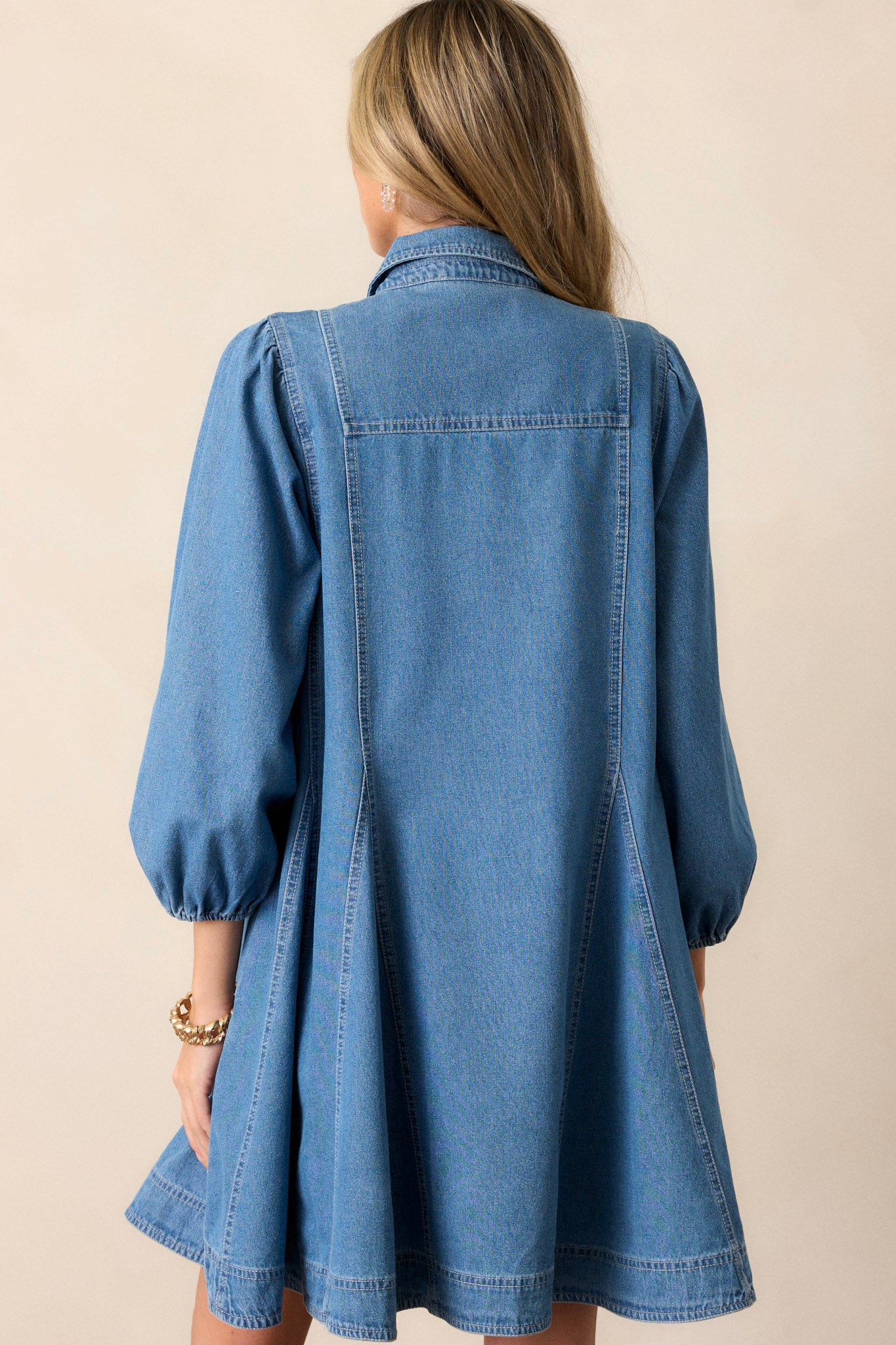 Back view of this medium wash denim dress that features a collared neckline, a functional button front, functional chest pockets, functional hip pockets, a relaxed fit and elastic cuffed three-fourth sleeves.