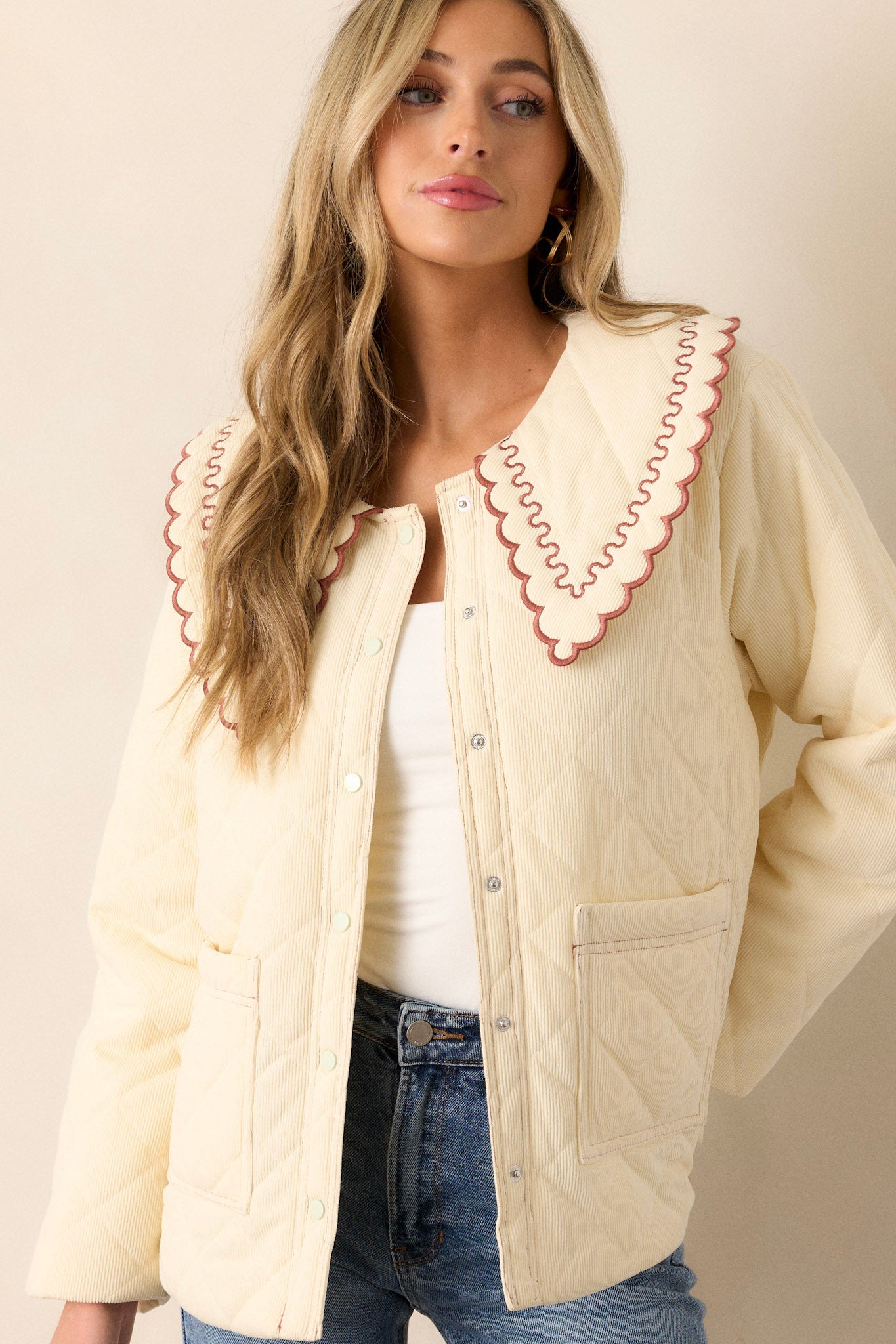 Front angled view of this champagne jacket that features a large collared neckline, a burnt orange ruffle trim on the collar, a functional button and snap front, functional hip pockets, and a soft quilted material.