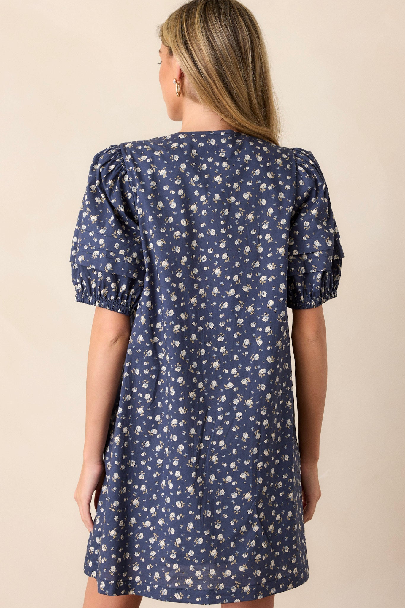 Back view of this navy dress that  features a v-neckline, pleats in the bust, a relaxed fit, a floral pattern and short puff sleeves.