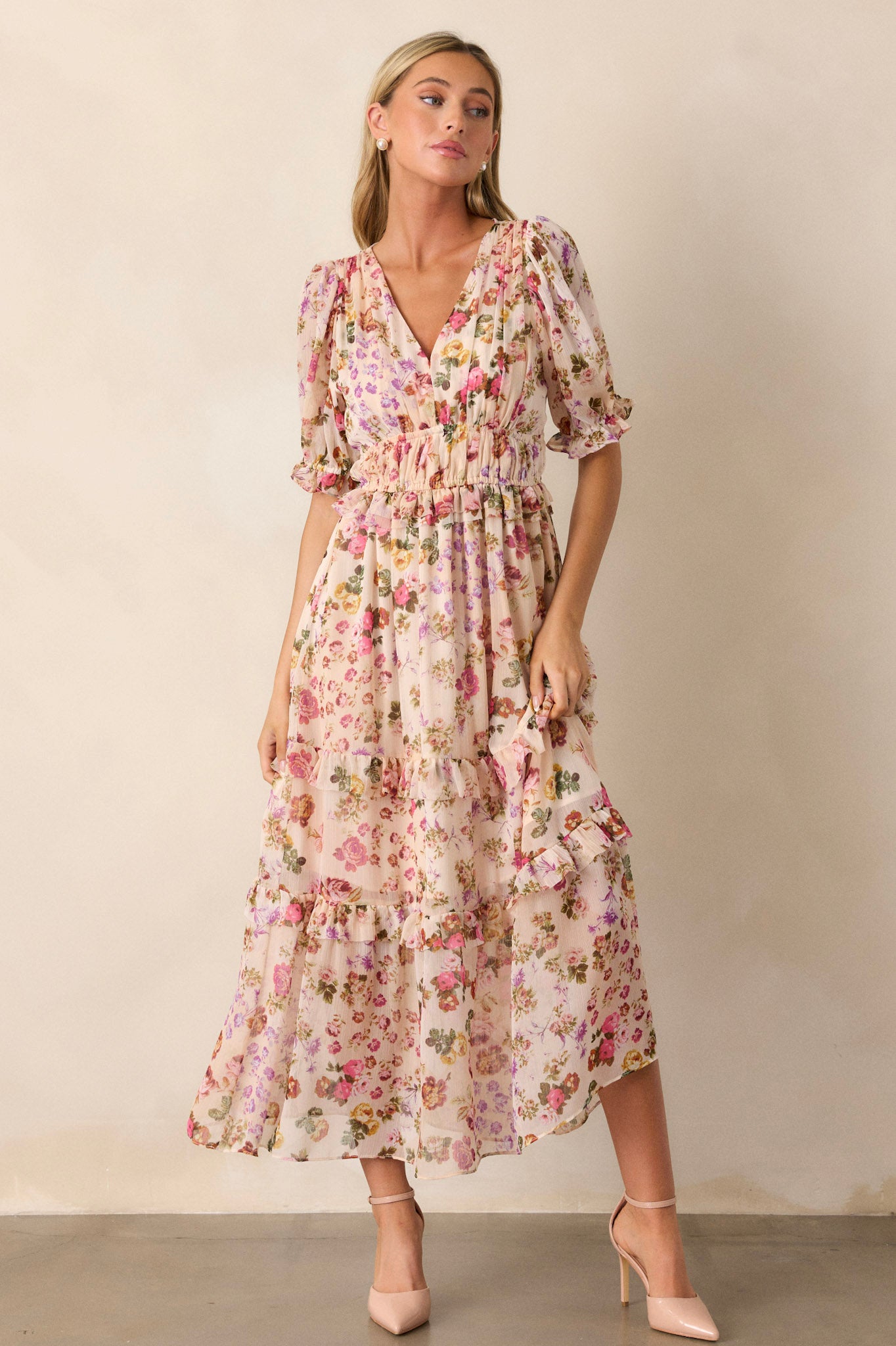 Full length view of this beige dress that features a v-neckline, two thin elastic waist bands, a tiered design, ruffled trim on the tiers, a pink floral design, a midi length and half length puff sleeves with elastic cuffs.