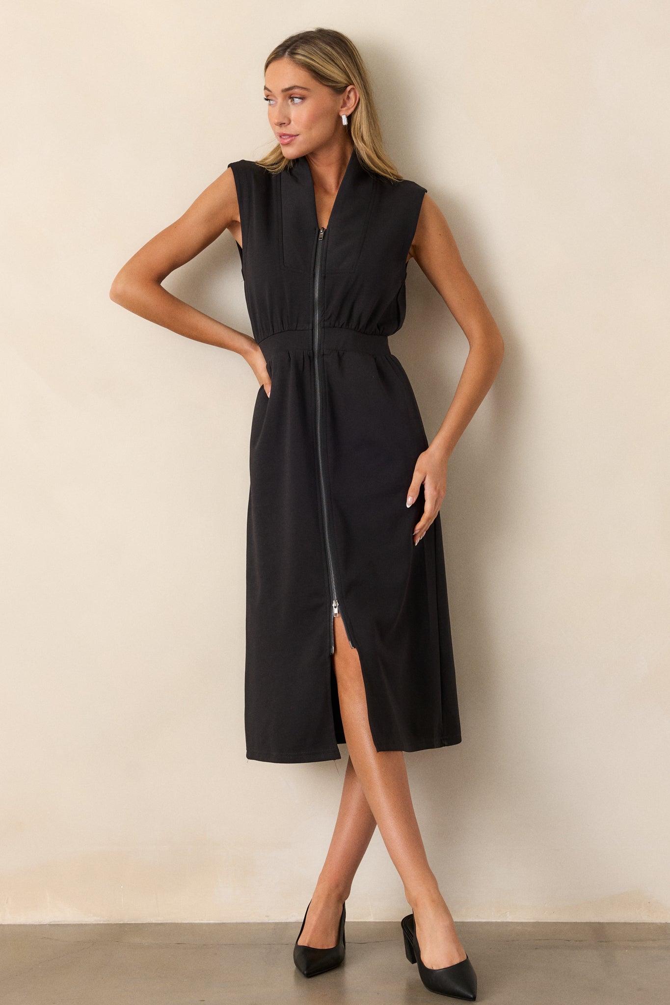 Front view of this black dress that features a v-neckline, a slight collar, a zipper front, functional hip pockets, a middle split in the hem and a sleeveless design.