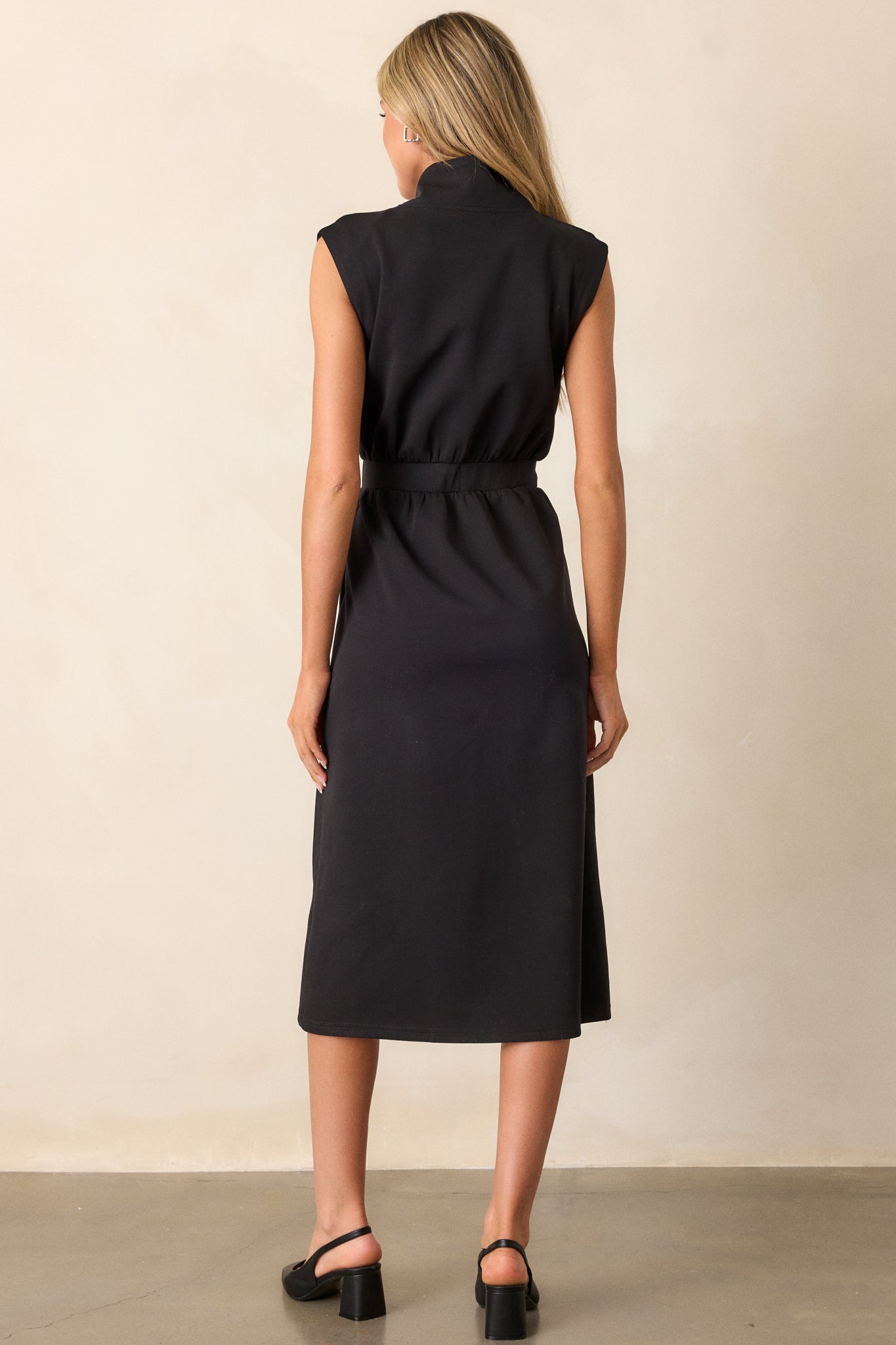 Back view of this black dress that features a v-neckline, a slight collar, a zipper front, functional hip pockets, a middle split in the hem and a sleeveless design.