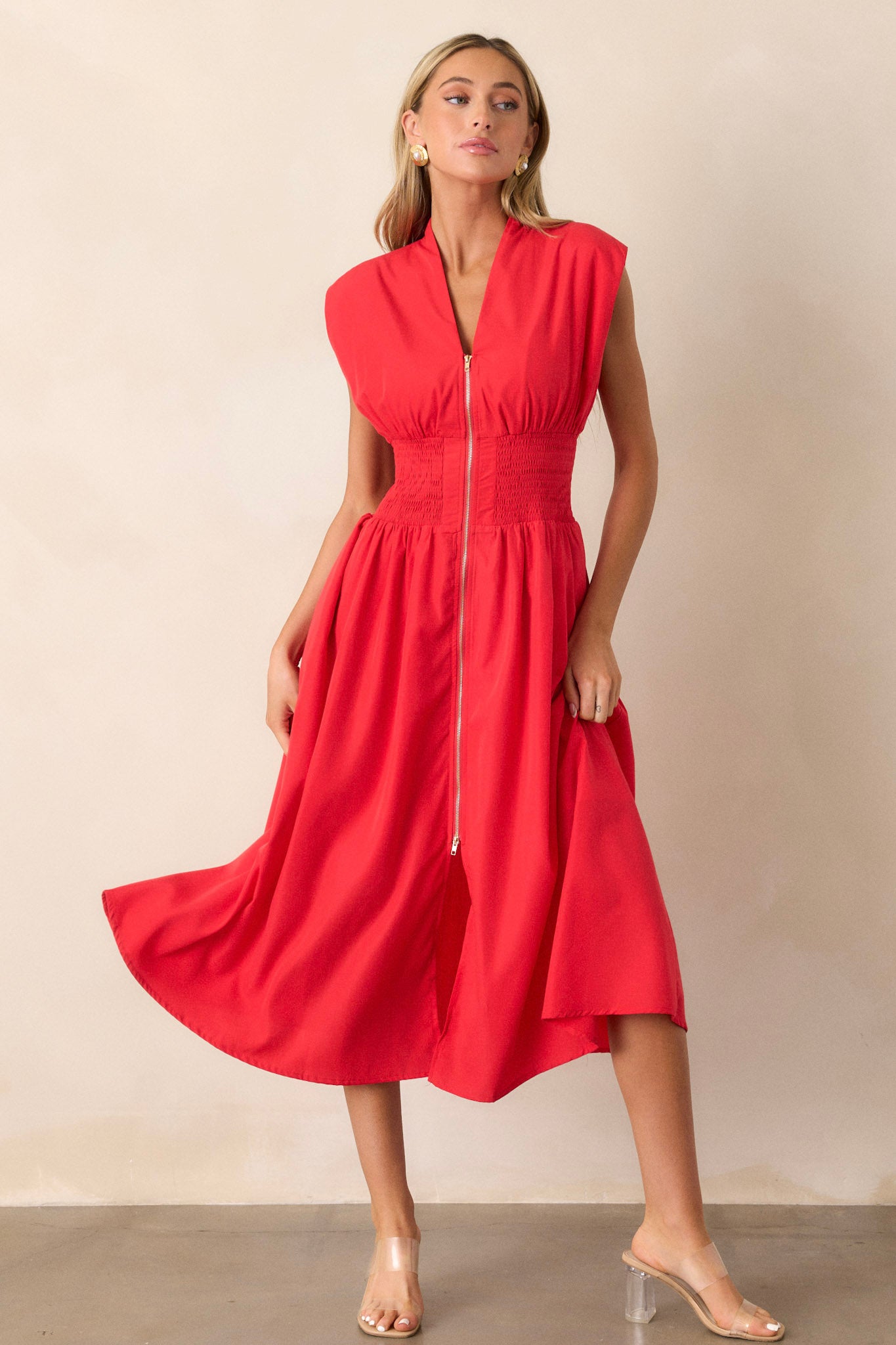 Full body view of this red midi dress that features a v-neckline, padded shoulders, a full zipper front, a fully smocked waist, functional hip pockets, and a front slit.