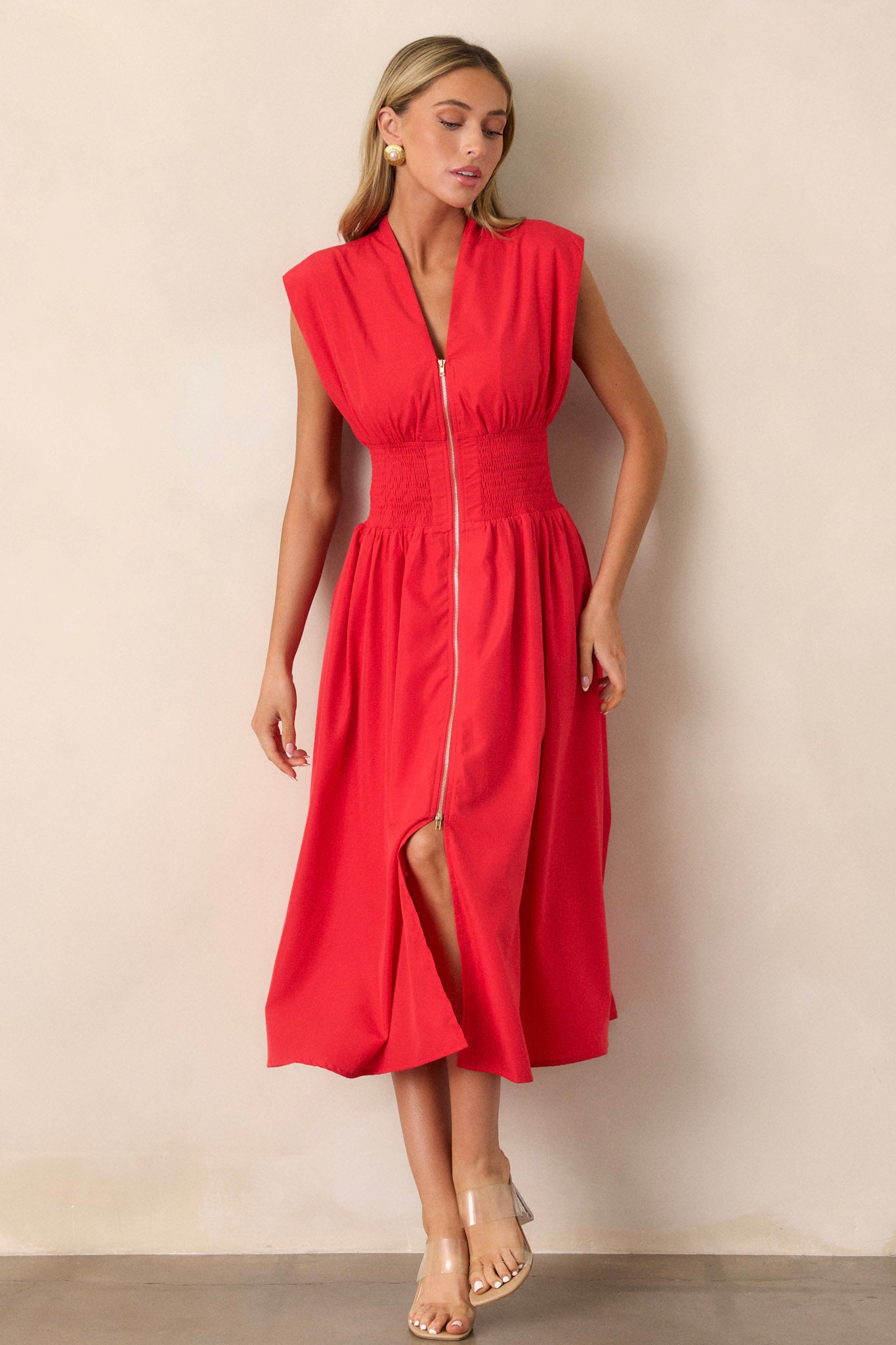 This red midi dress features a v-neckline, padded shoulders, a full zipper front, a fully smocked waist, functional hip pockets, and a front slit.