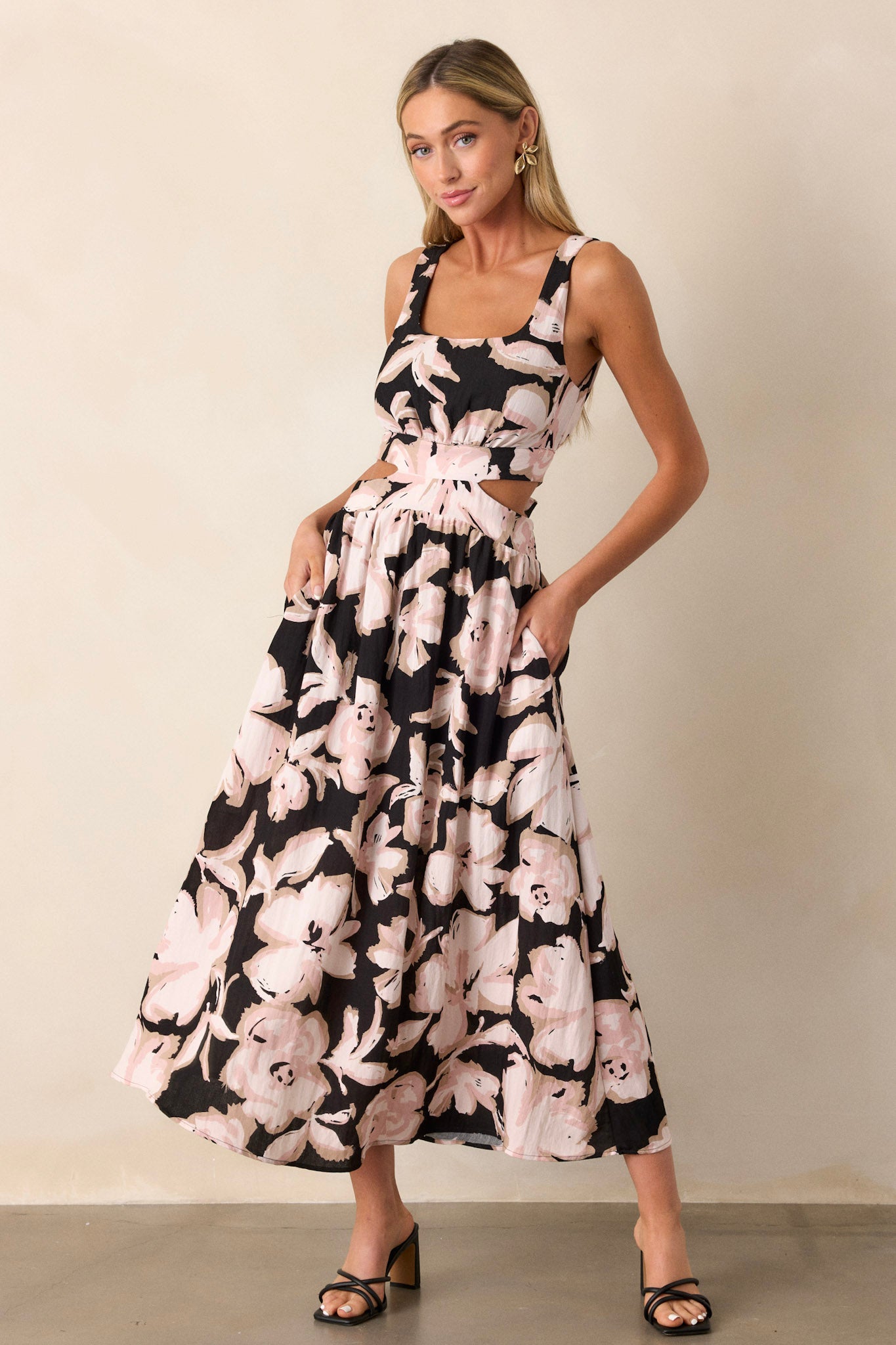 Angled front view of this black maxi dress that features a square neckline, thick straps, an elastic back insert, a self-tie back feature, waist cutouts with a smocked back insert, functional hip pockets, and an all over floral pattern.