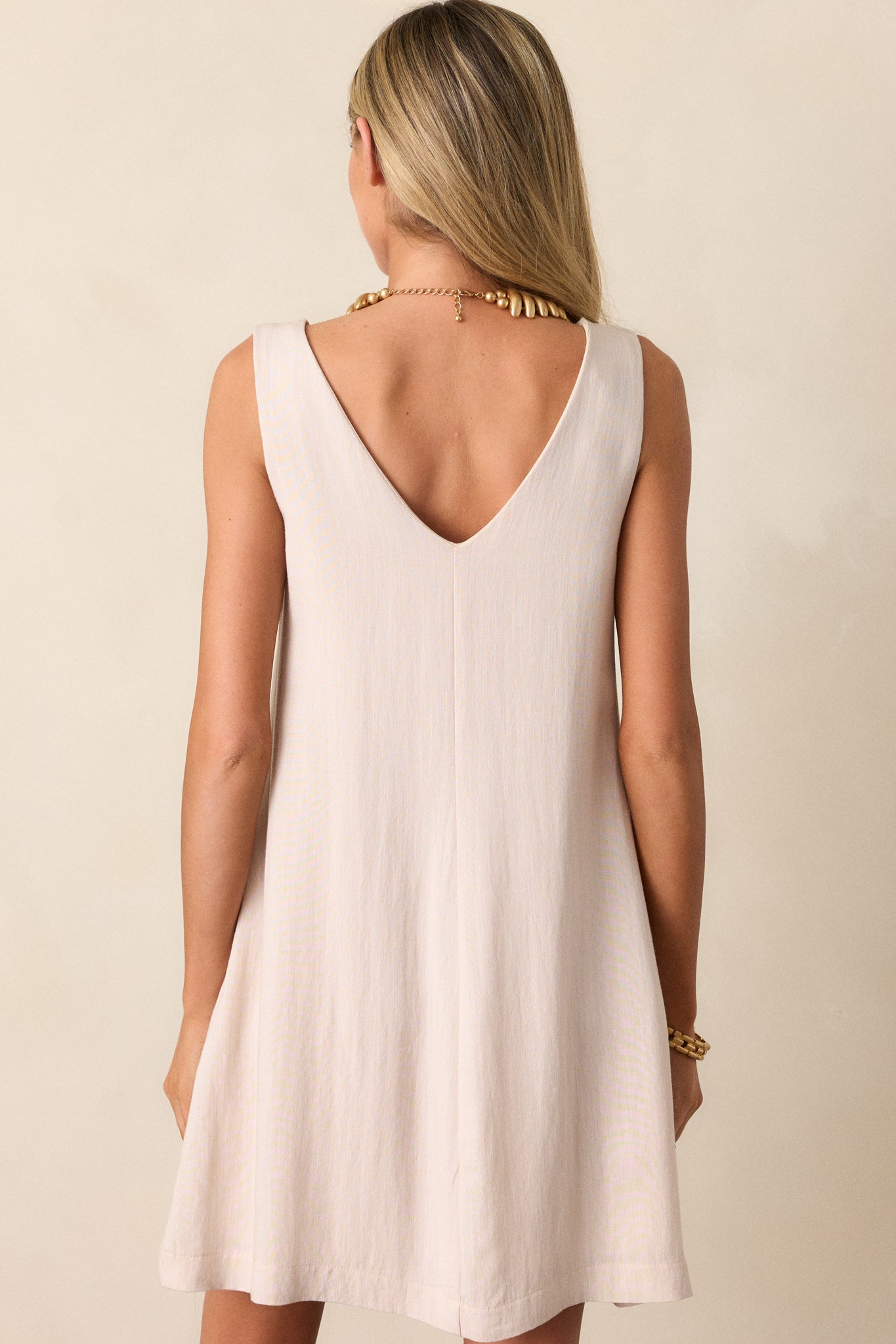 Back view of an ivory mini dress featuring a v-neckline, a sleeveless design, two functional hip pockets, and a shift silhouette.