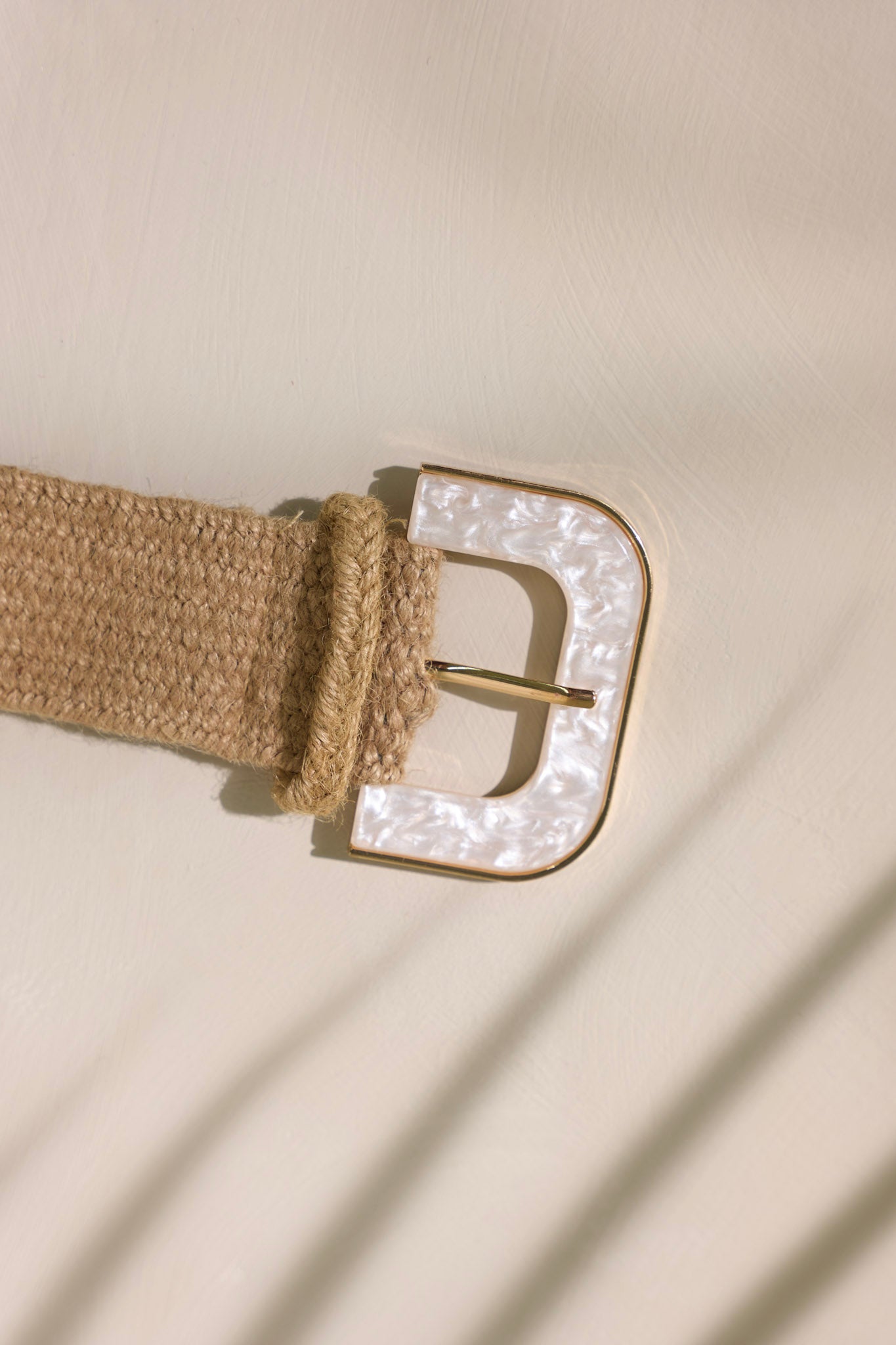 Close-up view of this tan belt that features a distinctive d-shaped buckle, an iridescent faux marble buckle with a gold trim and a woven material.