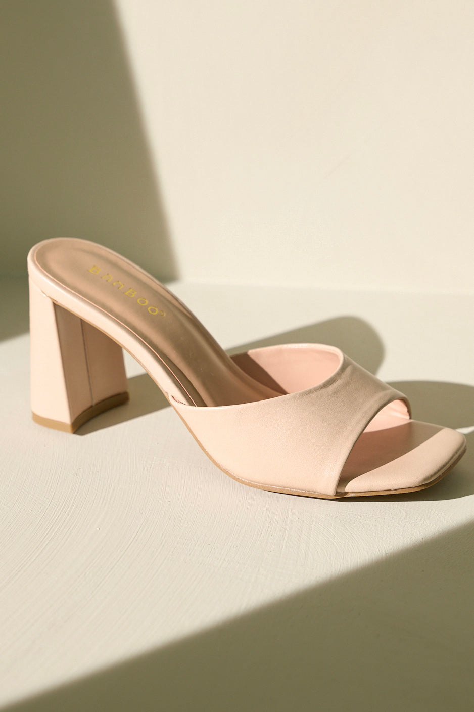 Front view of these beige heels that feature a square toe, a slip-on design, a strap over the top of the foot, and a thick block heel.