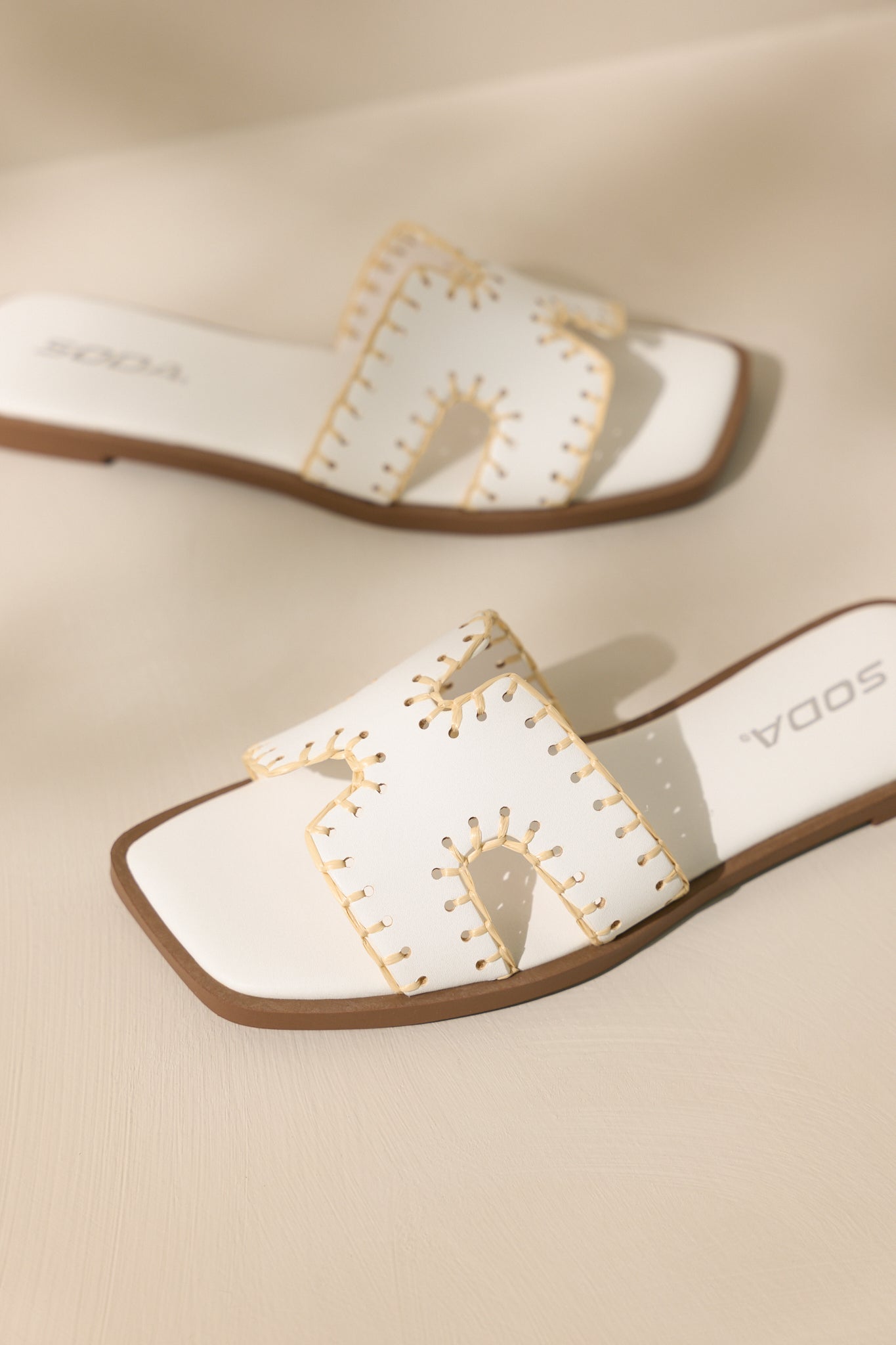 Detailed view of these white sandals that feature a square toe, a slip on design, a strap with cutouts over the top of the foot beige stitch detailing, and a high contrasting sole.