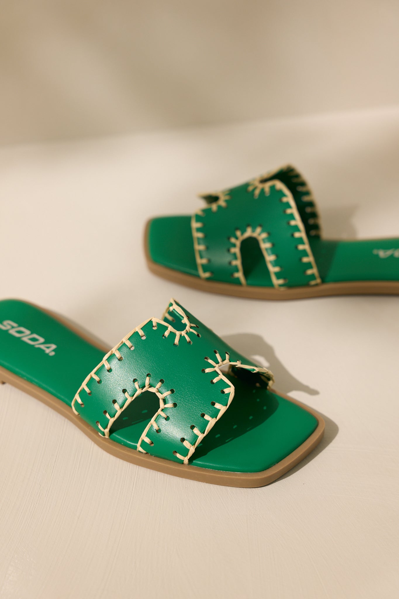 Close-up view of these green sandals that feature a square toe, a slip on design, a strap with cutouts over the top of the foot beige stitch detailing, and a high contrasting sole.
