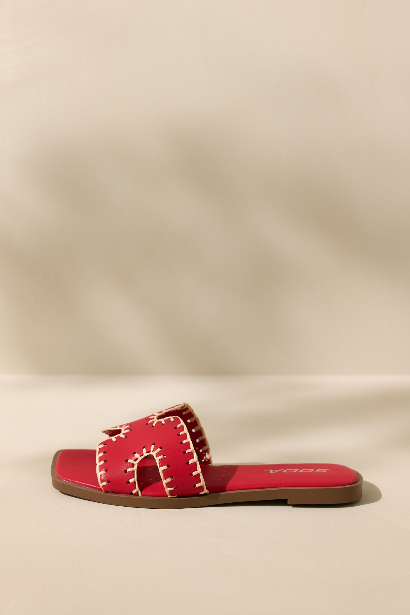 Side view of these red sandals that feature a square toe, a slip on design, a strap with cutouts over the top of the foot beige stitch detailing, and a high contrasting sole.