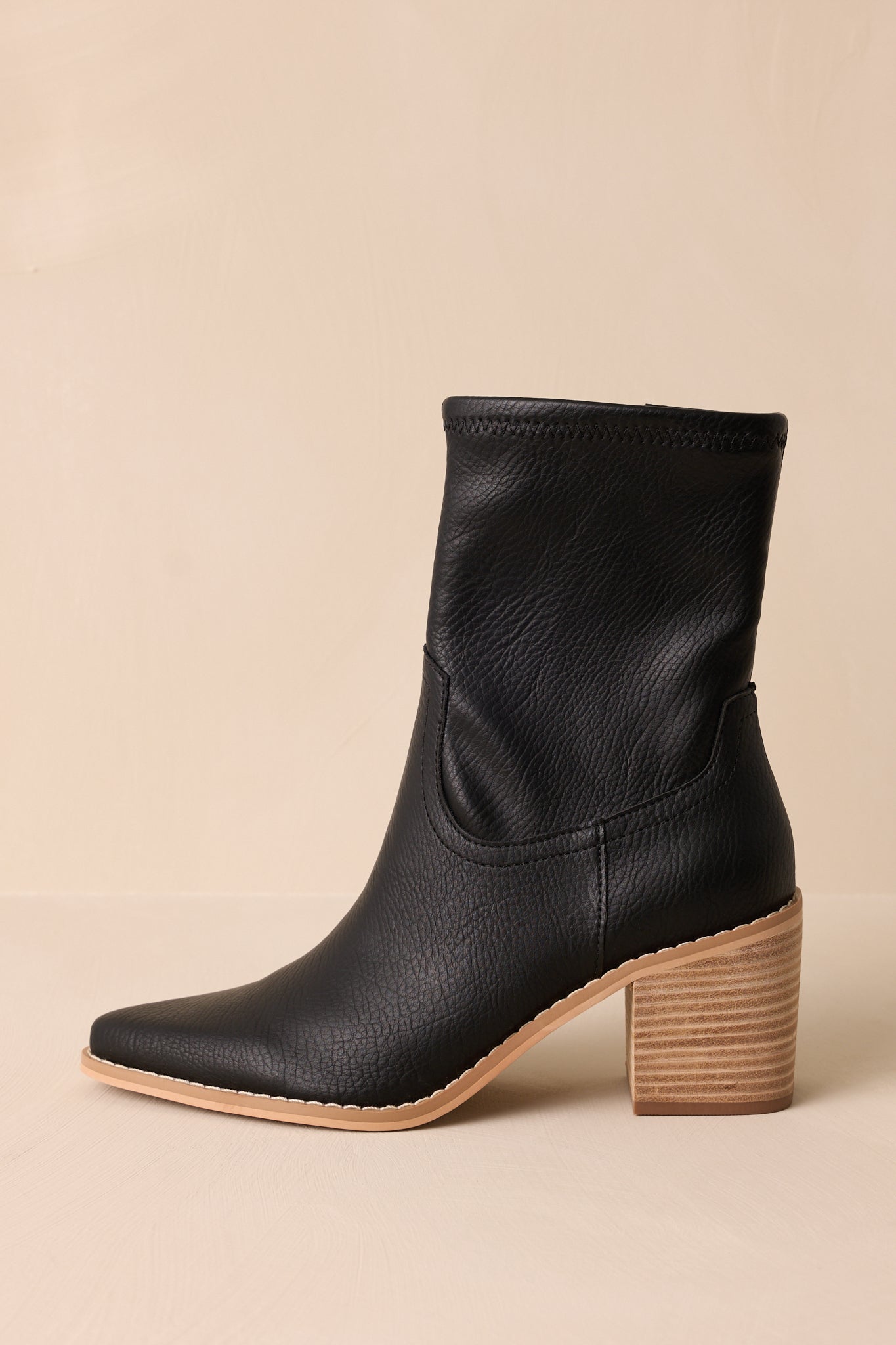 Side view of these black boots that feature a pointed toe, a platform heel, a discrete side zipper and a tan sole.