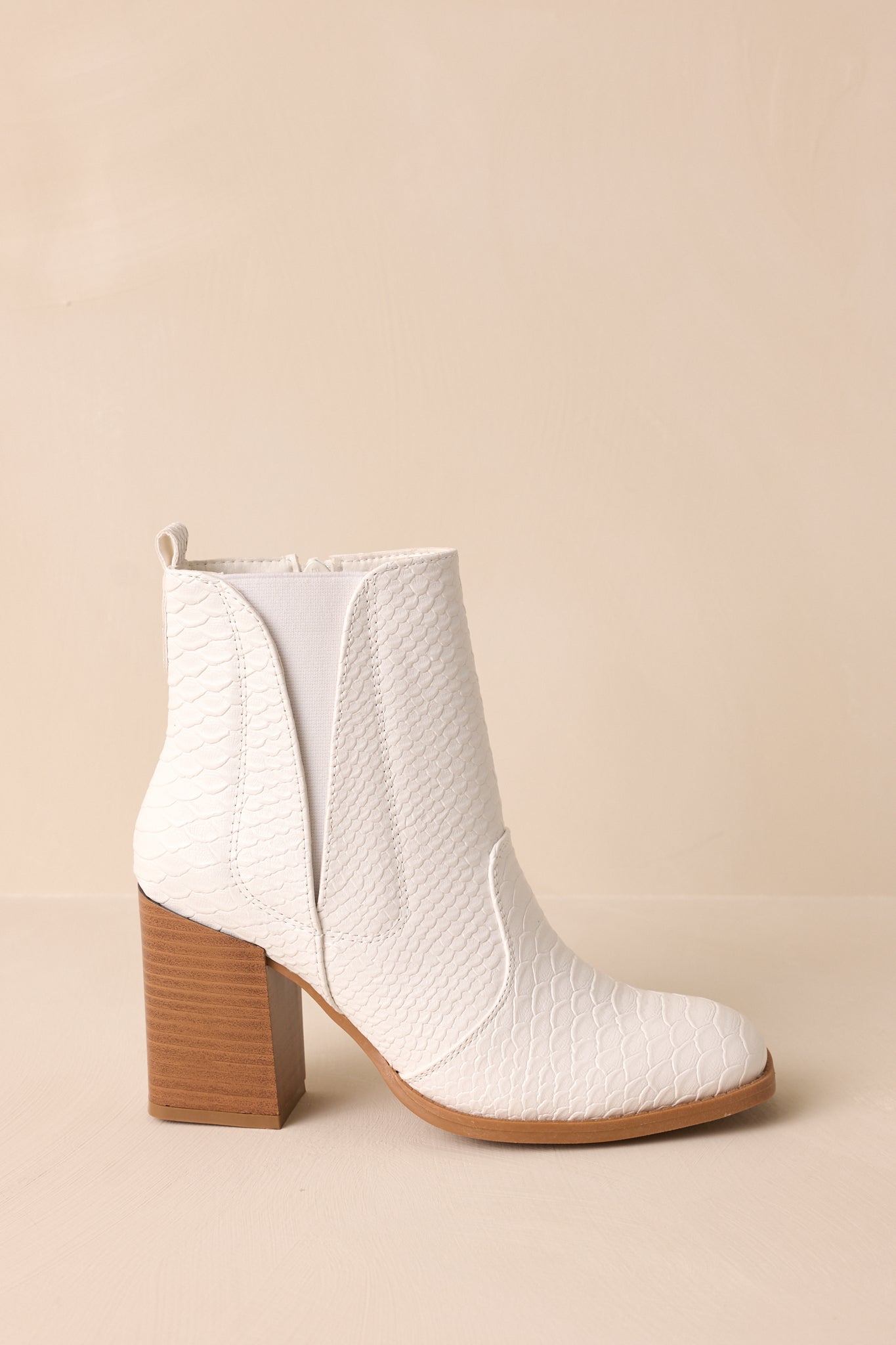 Close-up side view of these white ankle boots that feature a square toe, a platform heel, a snake skin texture, a side zipper and an elastic side insert.