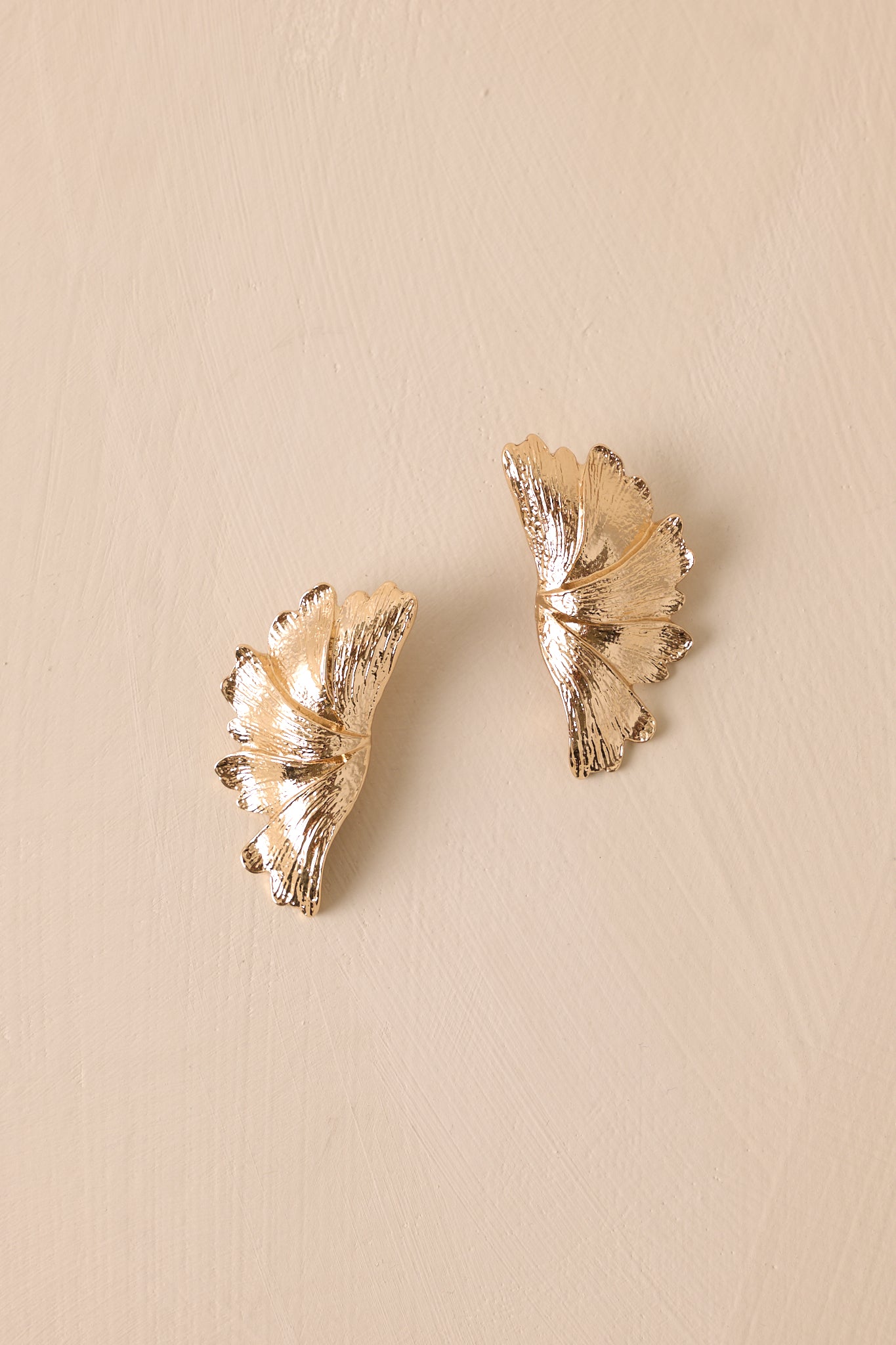 Overhead view of these textured gold earrings that feature gold hardware, a wing like shape, and secure post backings.