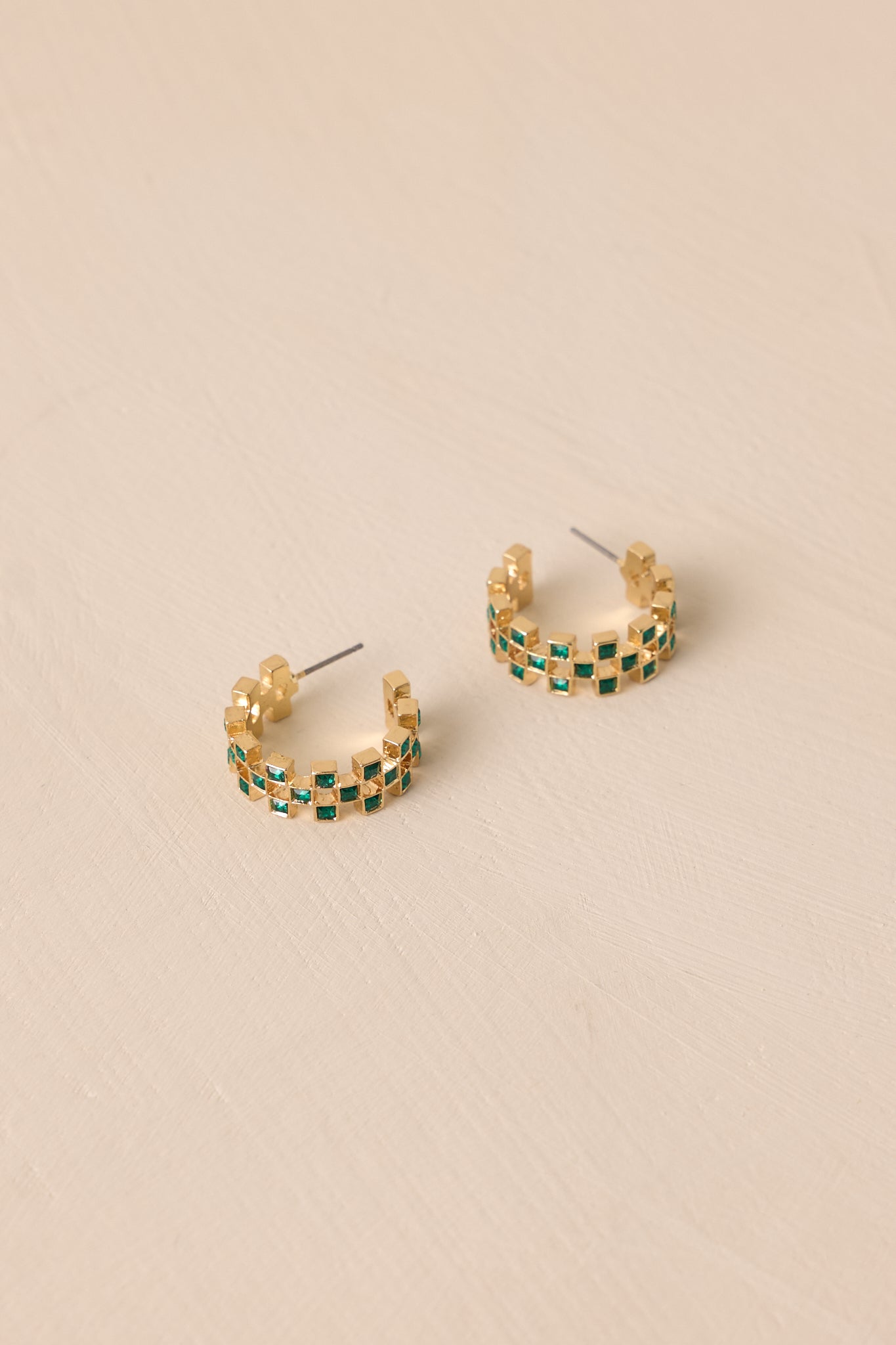 Two gold incomplete hoop earrings displayed on a neutral surface, featuring a checkered design with vibrant green rhinestones. The earrings have secure back posts, with a close-up view highlighting the texture and sparkle of the stones.