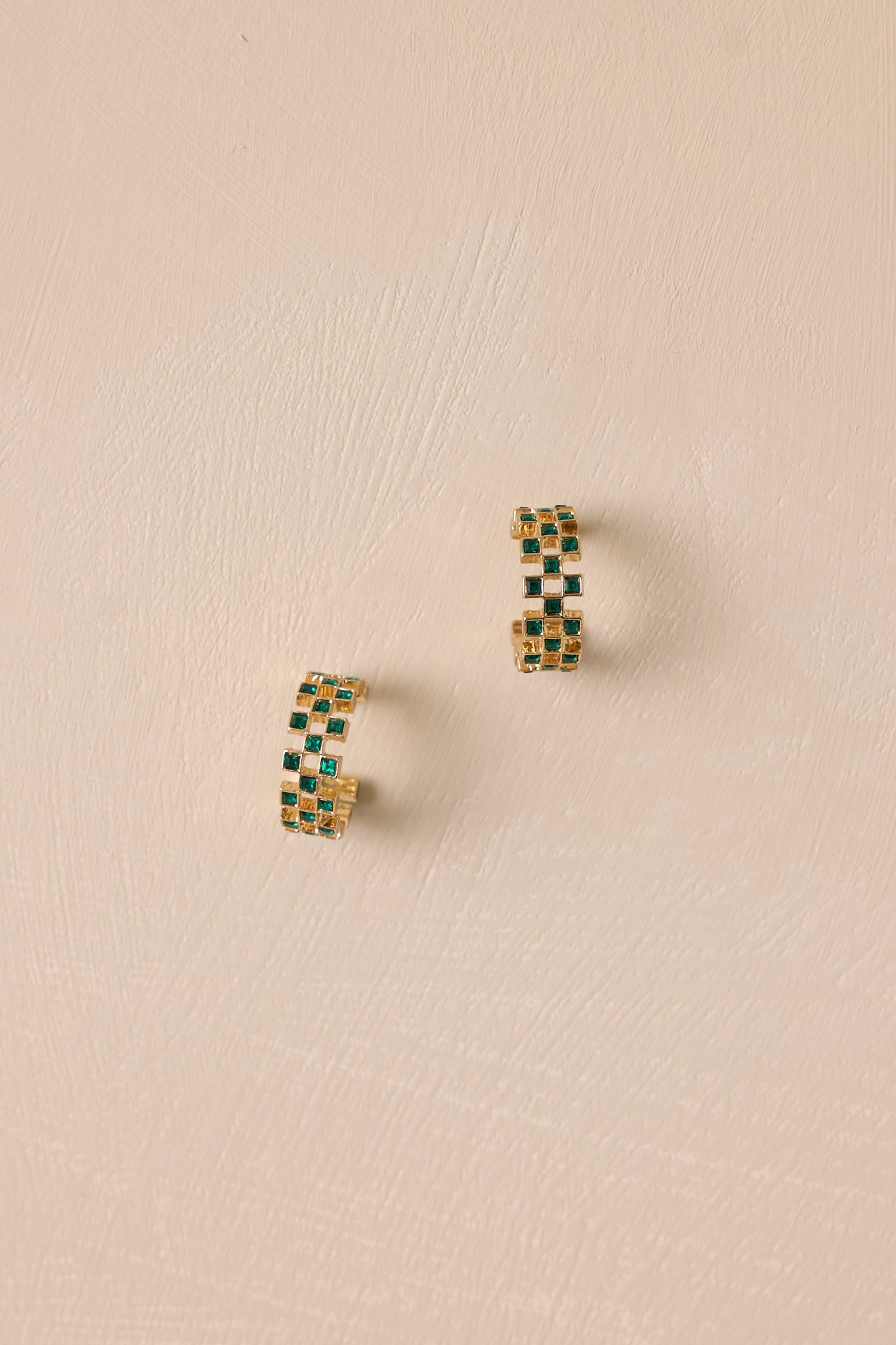 A pair of gold incomplete hoop earrings placed on a light-colored background. The earrings are adorned with small square-cut green rhinestones in a checkered arrangement and are designed with back postings for secure wear.