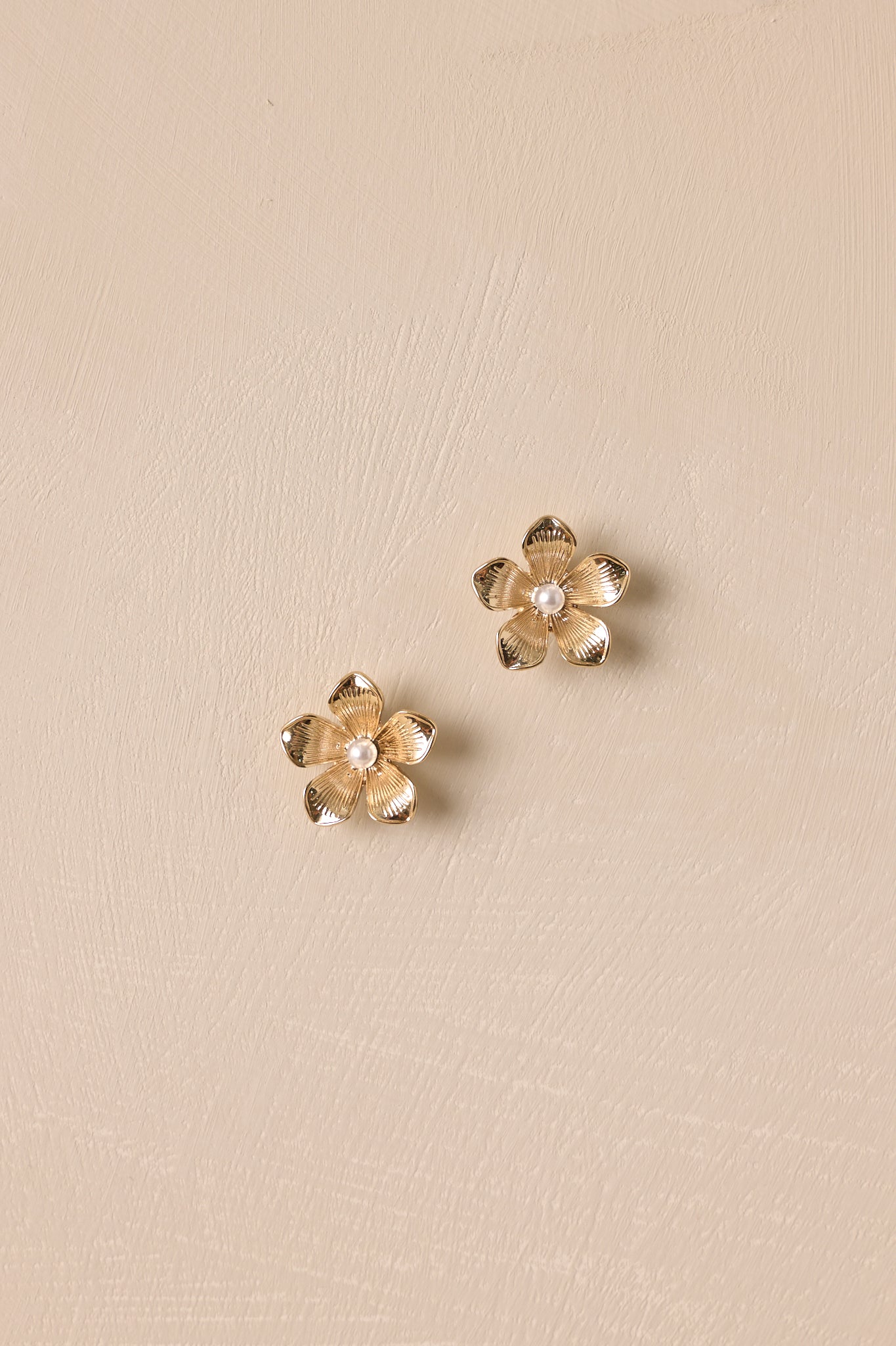 Overhead view of these gold earrings that feature a floral shape, a pearl center, and secure post backings.