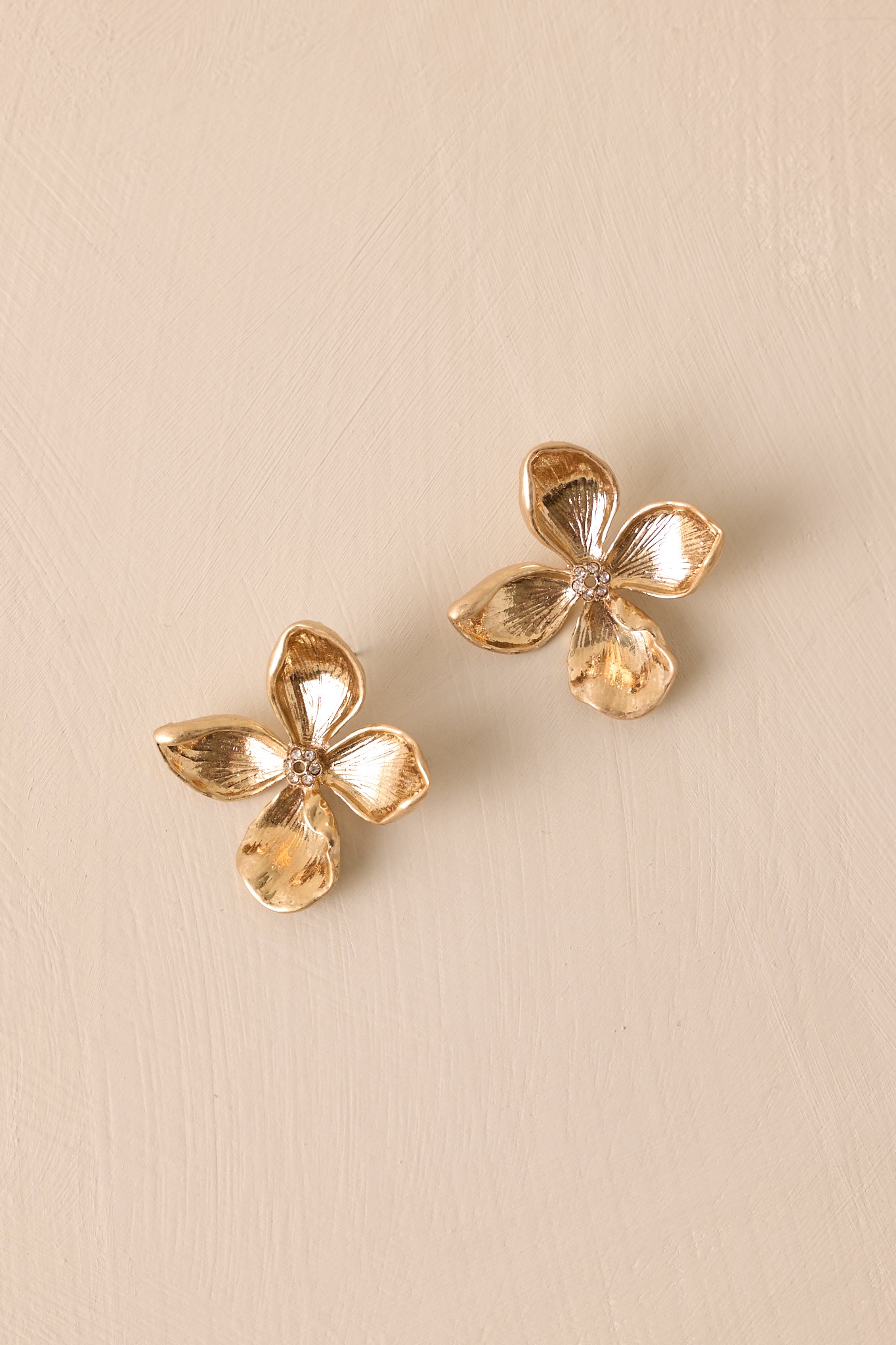 Over-head view of these gold flower earrings that feature a four petal flower, gold hardware, a rhinestone middle and secure back postings.