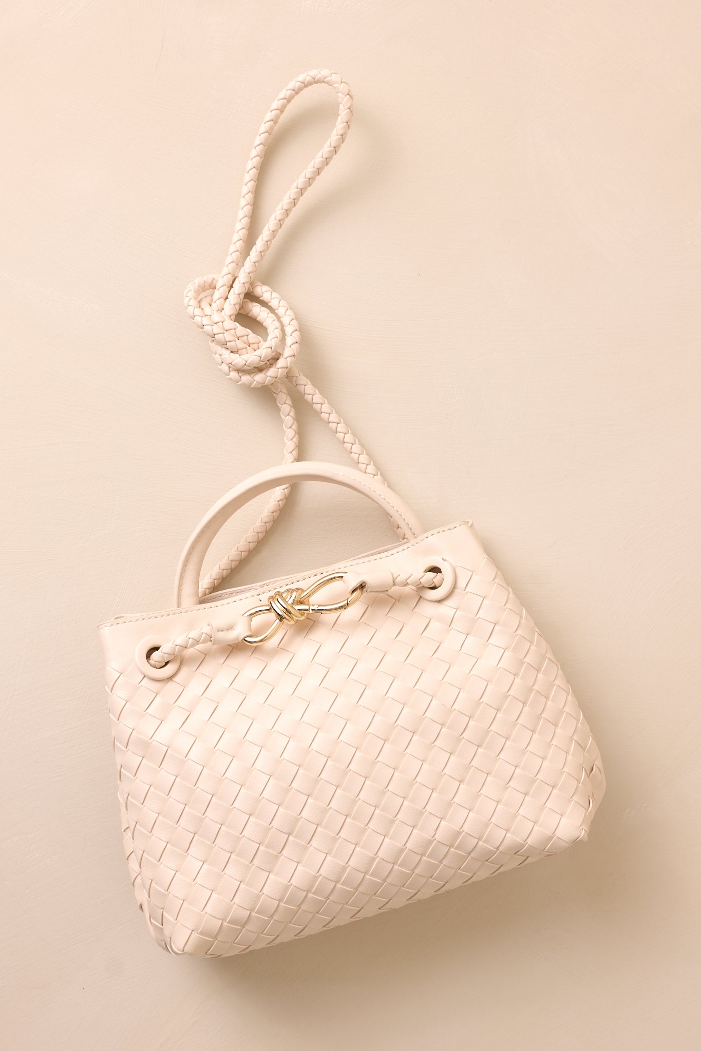 The ivory faux leather bag laid flat against the beige background, with the long removable woven strap draped across the bag.