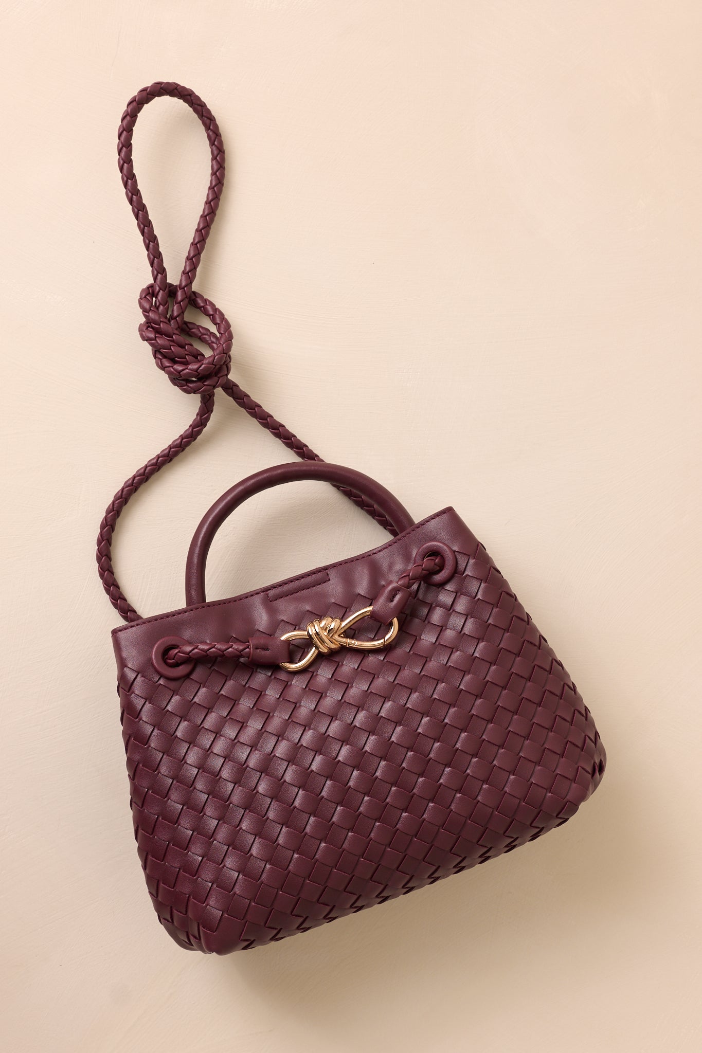 The wine faux leather bag laid flat against the beige background, with the long removable woven strap draped across the bag.