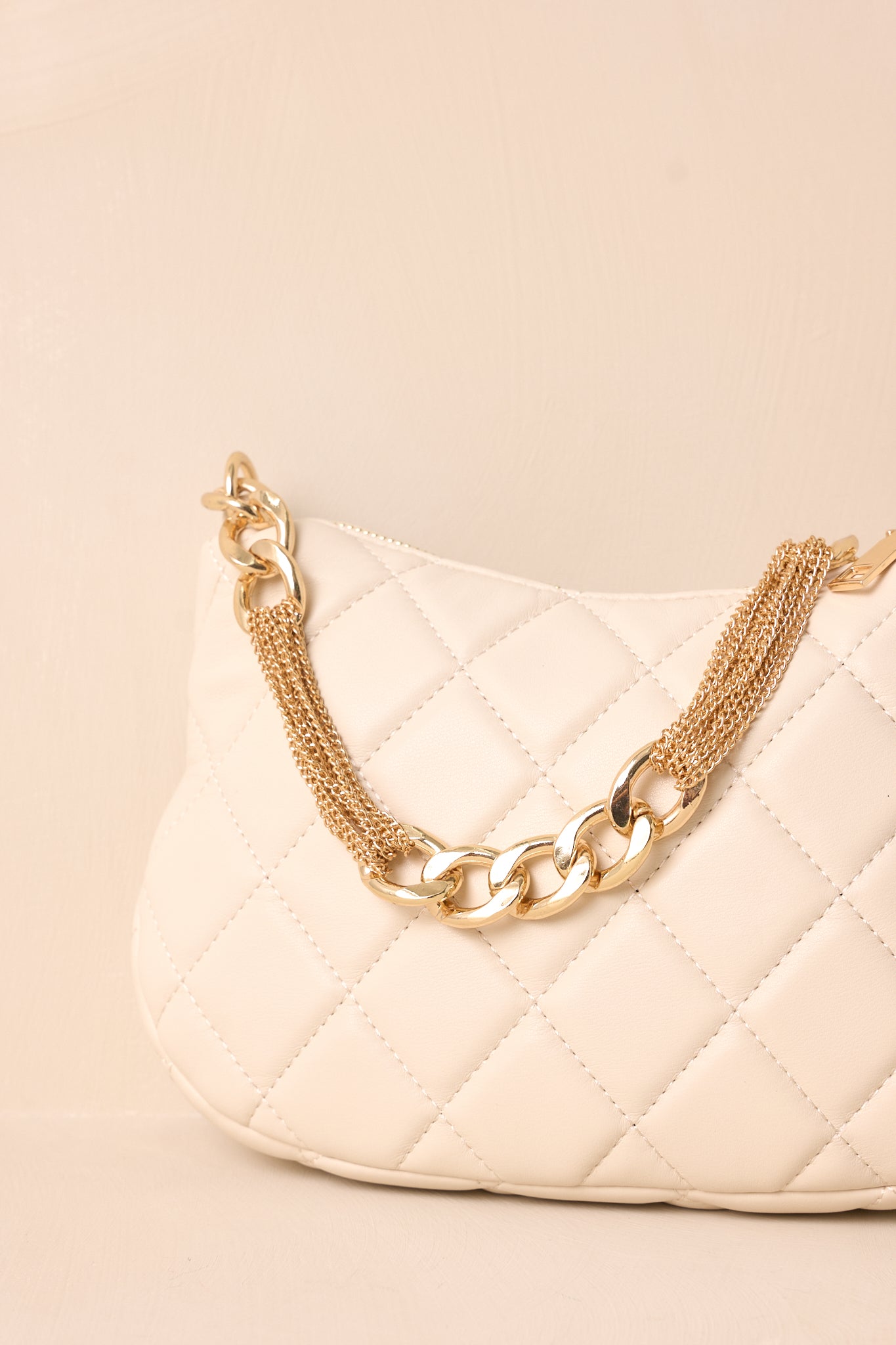 A detailed view of the ivory faux leather handbag, focusing on the fine quilted stitching, the gleaming gold zipper closure, and the attached thick gold chain handle.