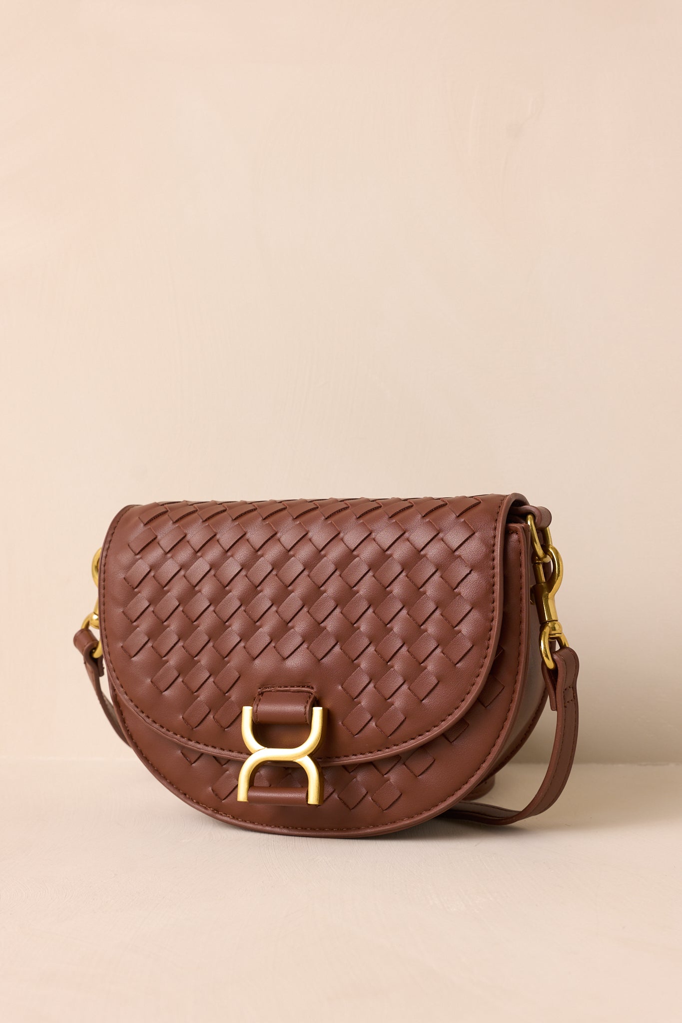 The brown handbag displayed upright against a beige background, showcasing its fold-over design, gold buckle snap closure, and woven faux leather material.