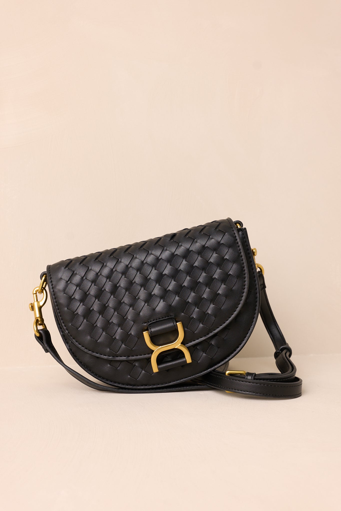 A side angle of the black handbag on a beige background, highlighting the solid black belt-style shoulder strap and the structured silhouette of the design.