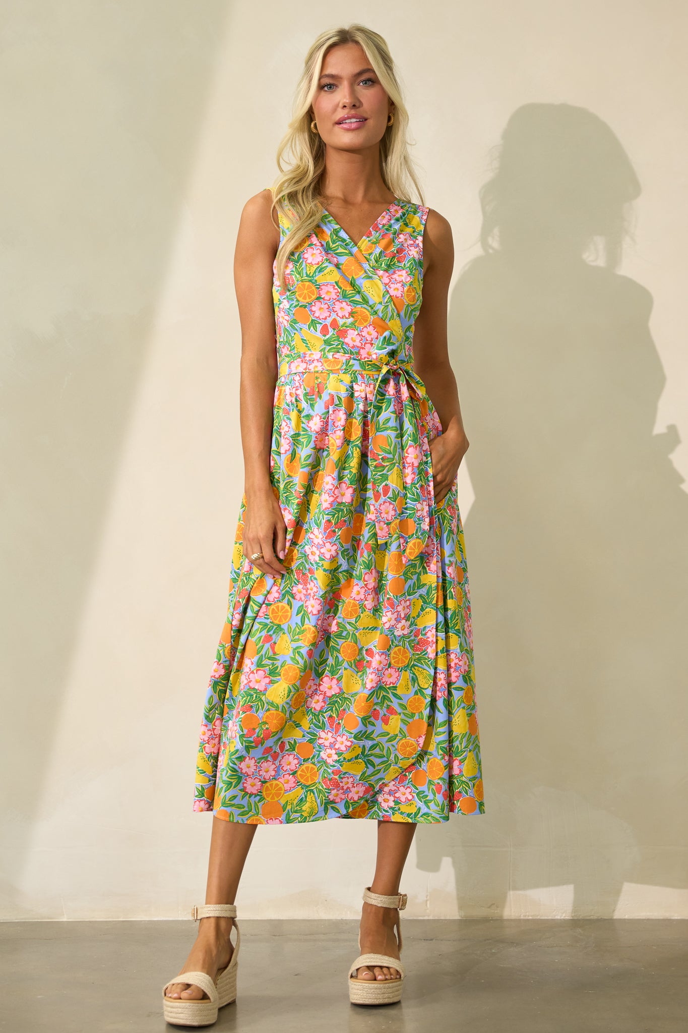 Full length view of a sky blue multi dress with a surplice neckline, a self-tie waist belt, functional hip pockets, a sleeveless design, and a flowing silhouette