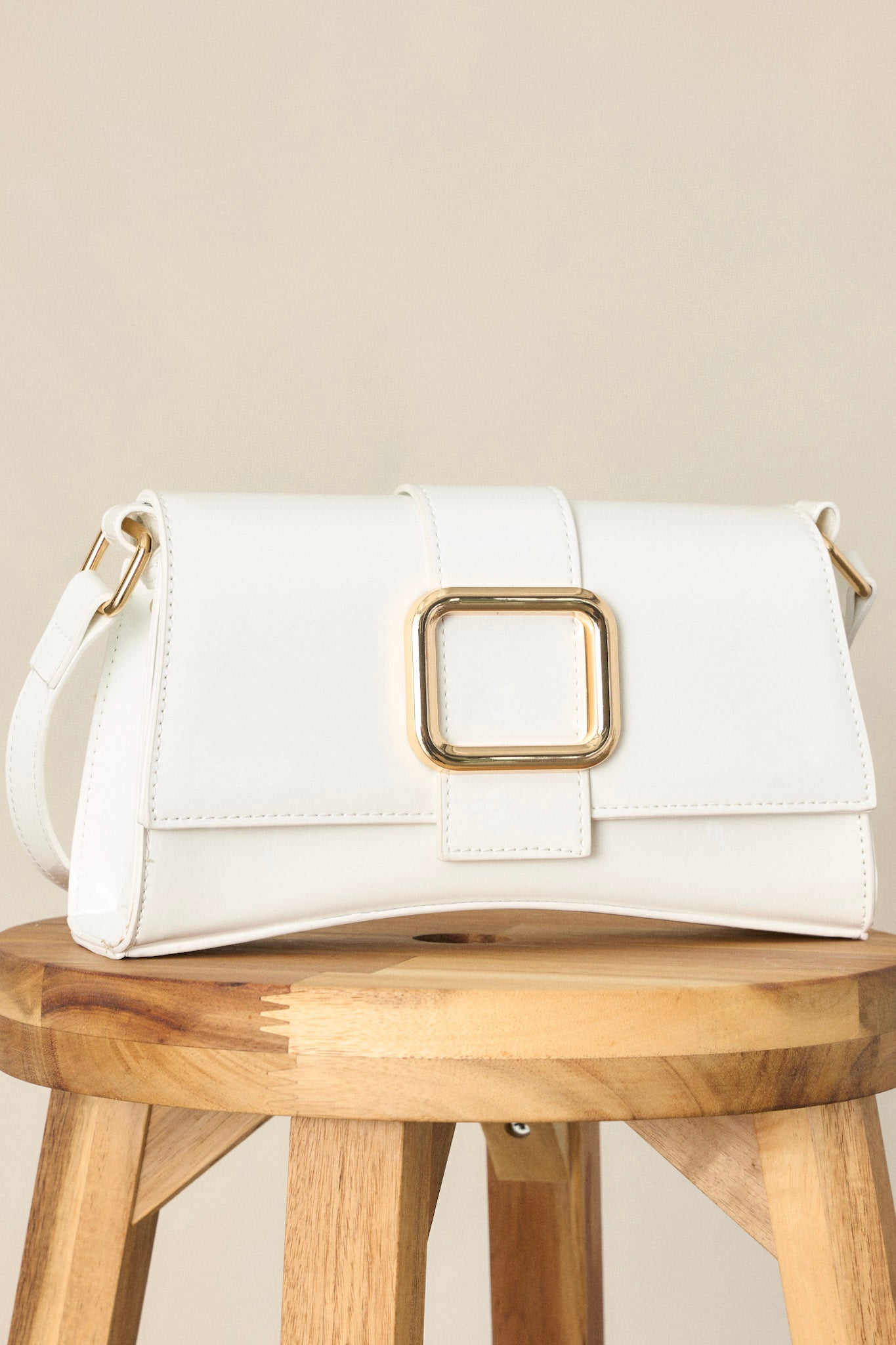 Close-up view of this white handbag featuring a gold rectangular buckle, two snap clasps, interchangeable straps and a glossy chic design.