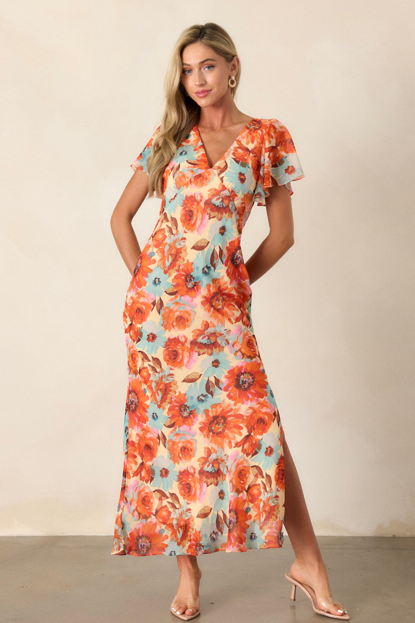 Action shot of an orange floral dress displaying the flow and movement, highlighting the v-neckline, floral print design, leg slit on the left leg, elastic waist insert, and sheer short sleeves